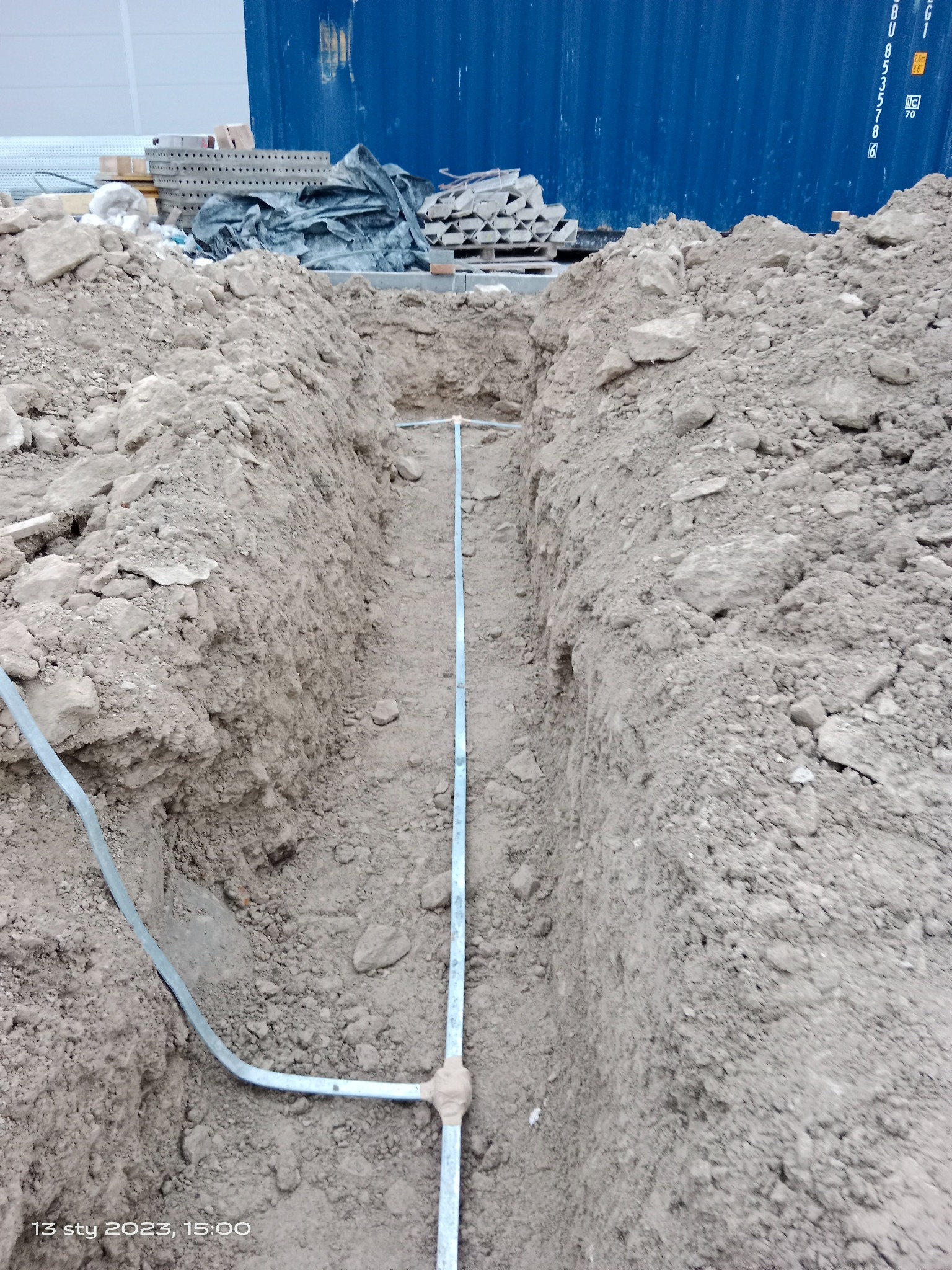 Wykop w ziemi z ułożonymi rurami, widoczne łączenia. W tle niebieski kontener i materiały budowlane.