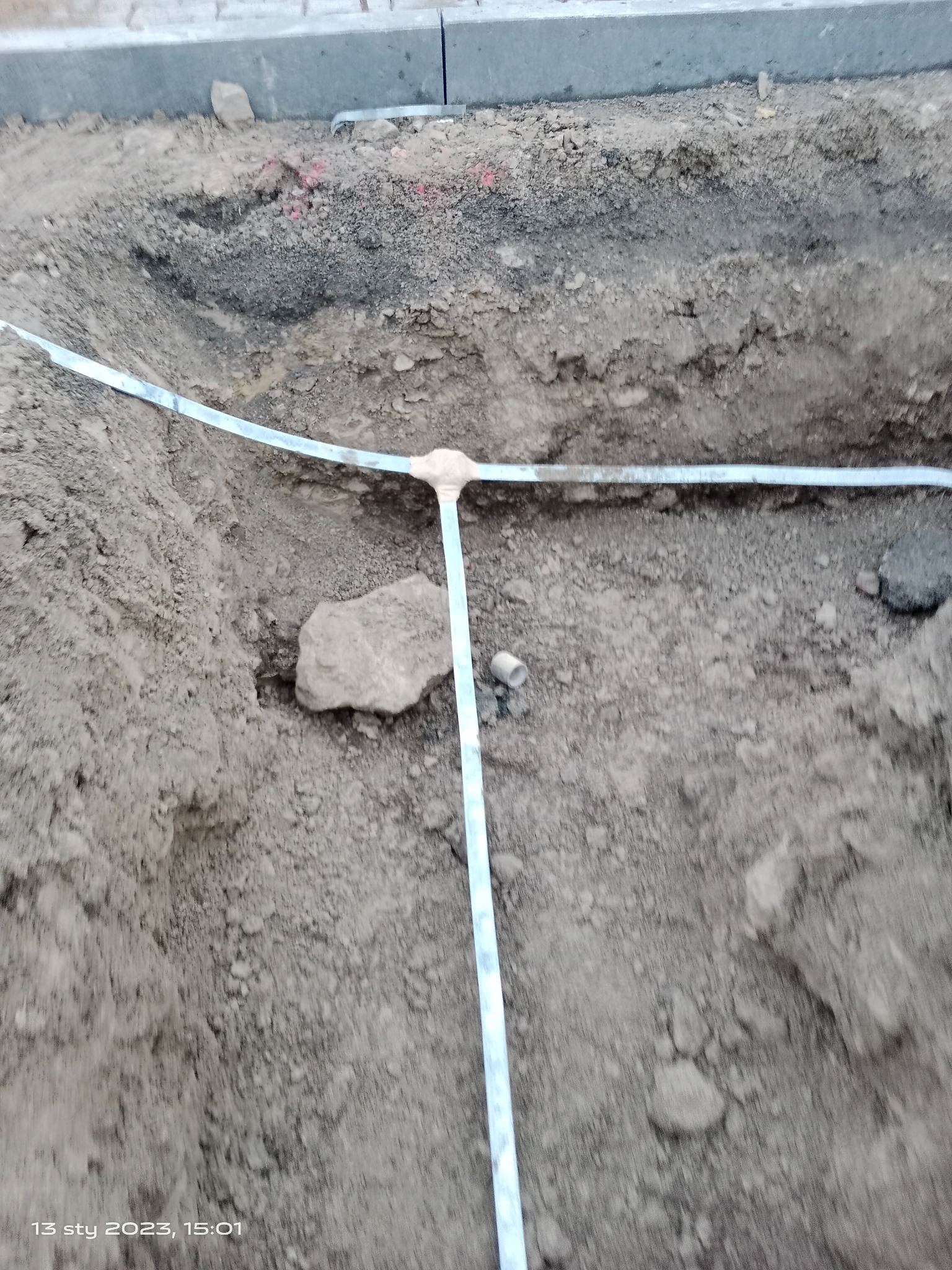 Wykop w ziemi z ułożoną taśmą odgromową w kształcie litery T, widoczne elementy instalacji uziemiającej.