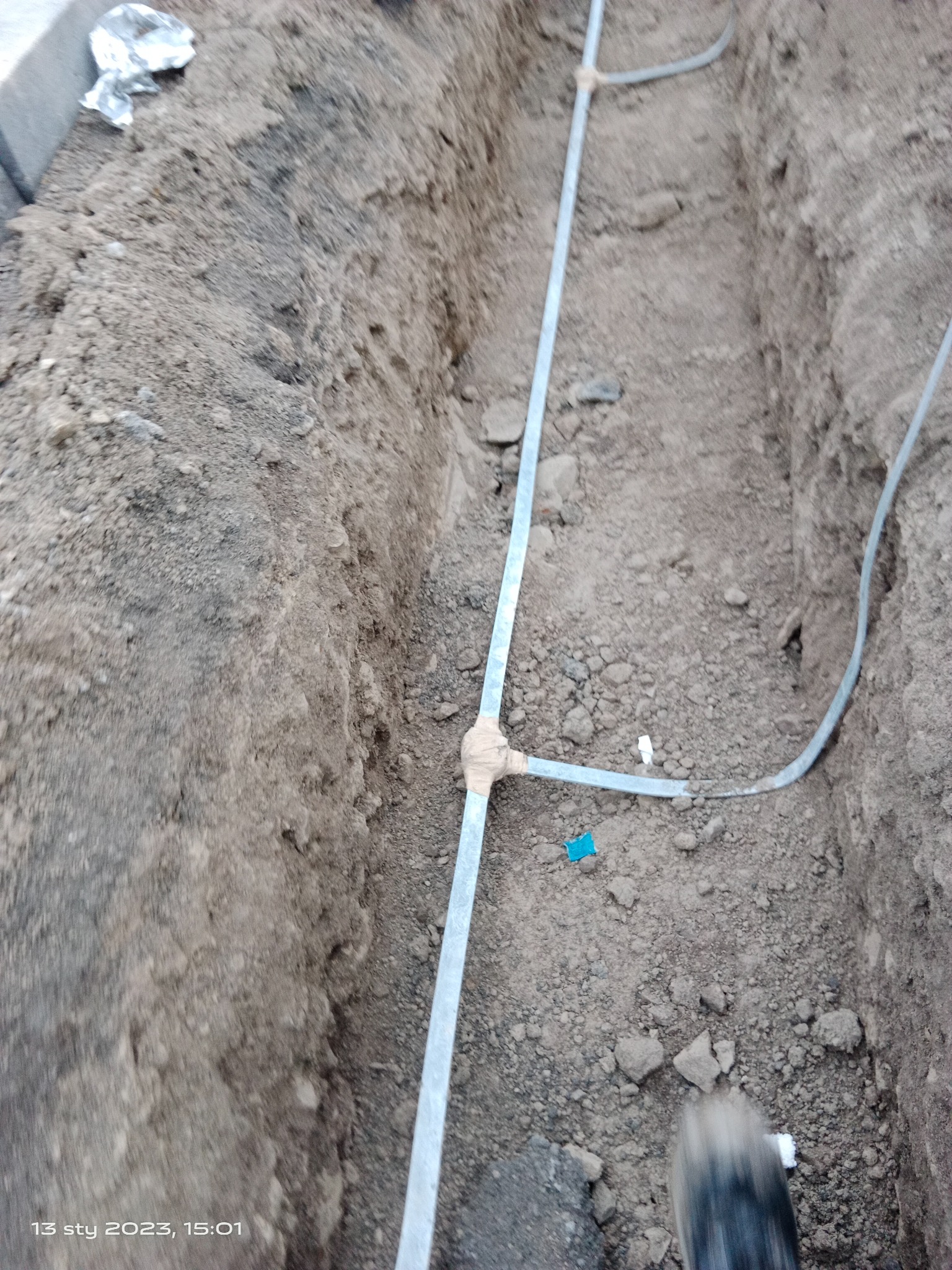 Wykop w ziemi z ułożonymi przewodami w metalowych rurkach, połączonymi taśmą izolacyjną, fragment buta w prawym dolnym rogu.