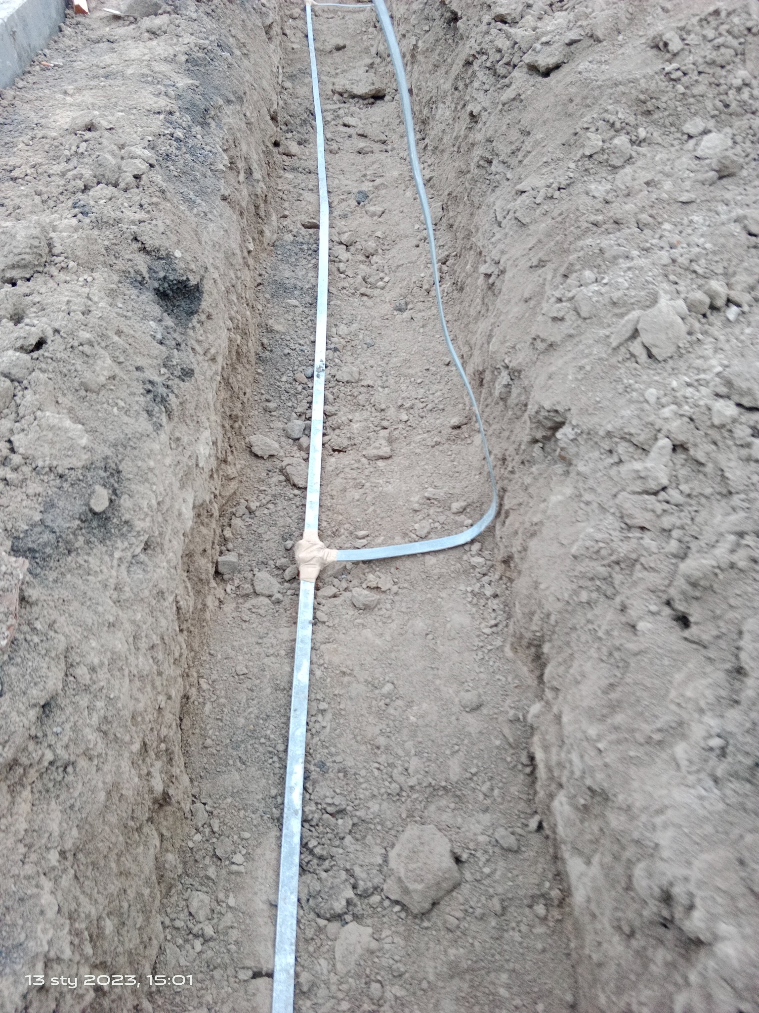 Wykop w ziemi z ułożoną metalową taśmą uziemiającą, zabezpieczoną taśmą izolacyjną w miejscu łączenia, widok z góry.