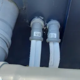 Szare rury kanalizacyjne z izolacją termiczną, przymocowane do ciemnej powierzchni, widoczne etykiety producenta 'Kolano HTplus 50/30' i 'Kolano HTplus 50188', pajęczyny w narożnikach.