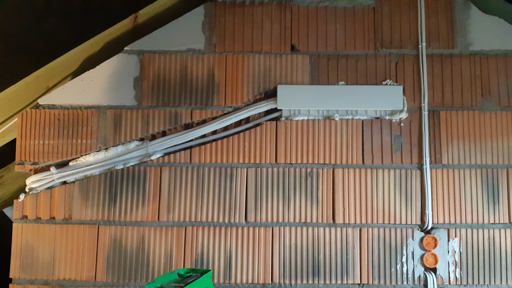 Instalacja przewodów w bruzdach ceglanej ściany, zabezpieczonych pianką montażową i plastikową listwą maskującą, widoczne puszki elektryczne.
