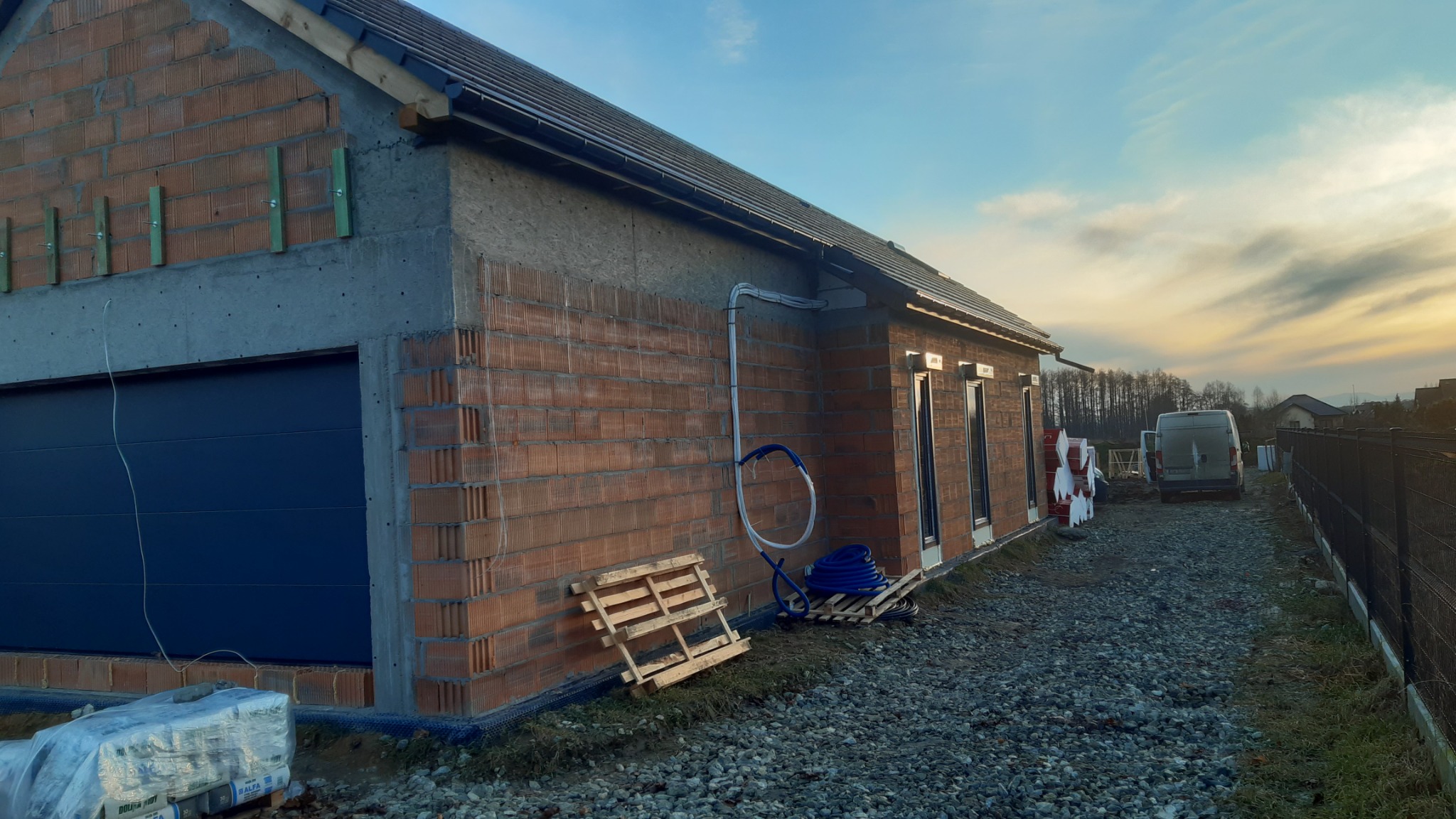 Surowy stan budynku jednorodzinnego z cegły, widoczna brama garażowa, instalacja elektryczna na elewacji oraz przygotowane rury do instalacji na zewnątrz, palety i materiały budowlane obok budynku,...