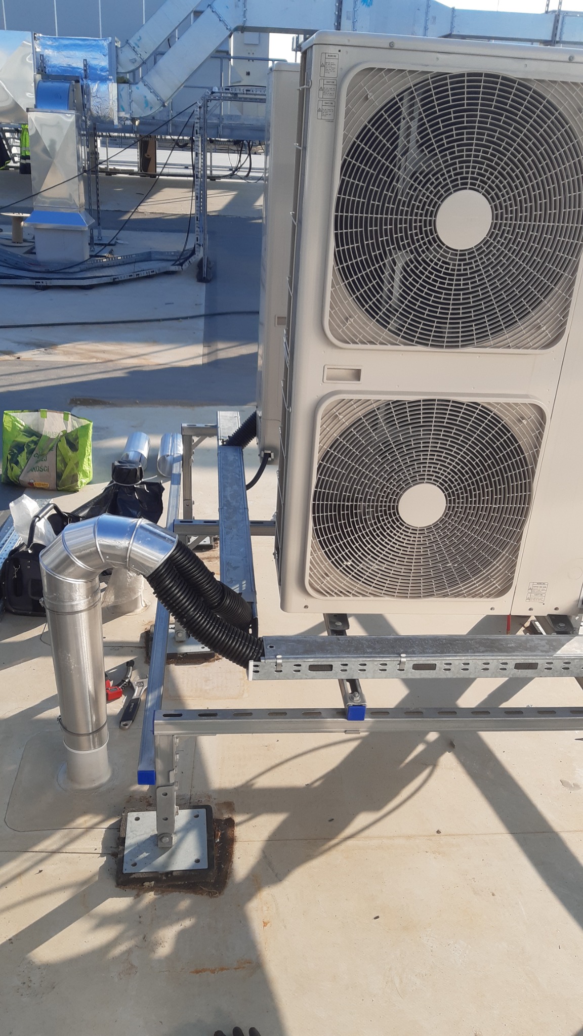 Dwa moduły klimatyzacyjne na dachu budynku, połączone metalowymi wspornikami i rurami wentylacyjnymi, widoczne narzędzia i torba robocza w tle.