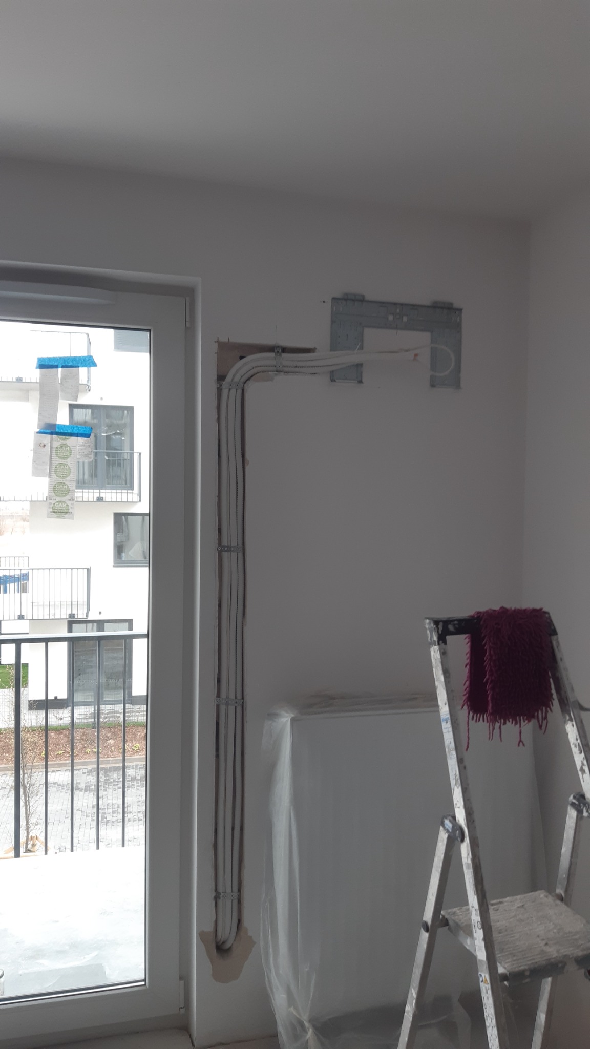 Przygotowanie instalacji pod klimatyzator w Ząbkowicach Śląskich: widoczne rury i przewody zamocowane do ściany, zabezpieczony mebel folią oraz drabina z bordowym pledem.