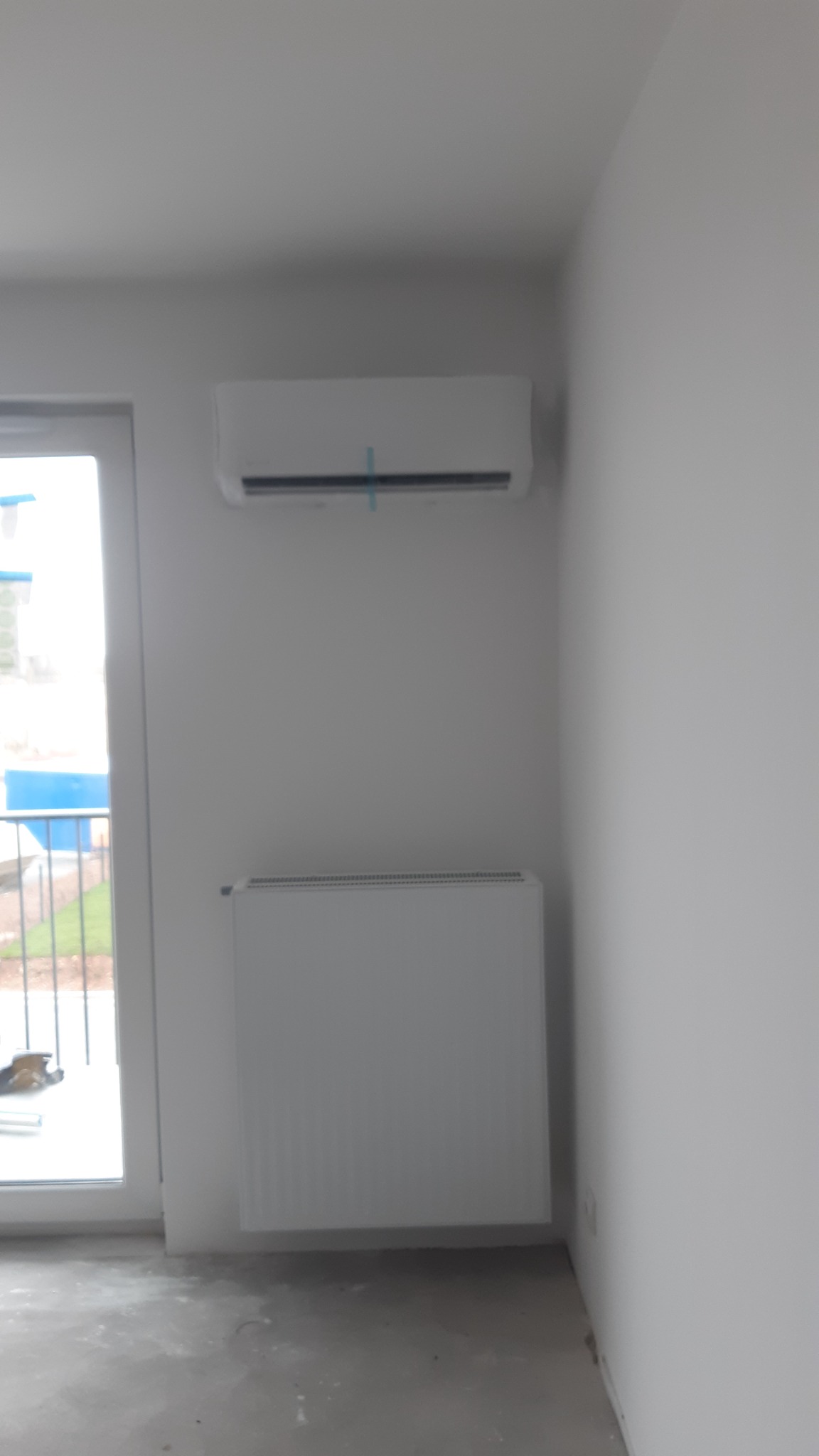 Biała jednostka wewnętrzna klimatyzacji zamontowana na ścianie w pokoju z grzejnikiem, widoczne zabezpieczenia montażowe na klimatyzatorze, surowy stan wykończenia podłogi.