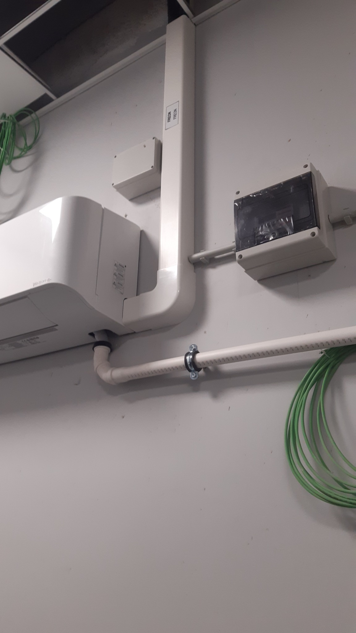 Biała jednostka wewnętrzna klimatyzacji z podłączonymi rurami i przewodami elektrycznymi na jasnej ścianie w biurze, widoczne elementy instalacji elektrycznej i wentylacyjnej.