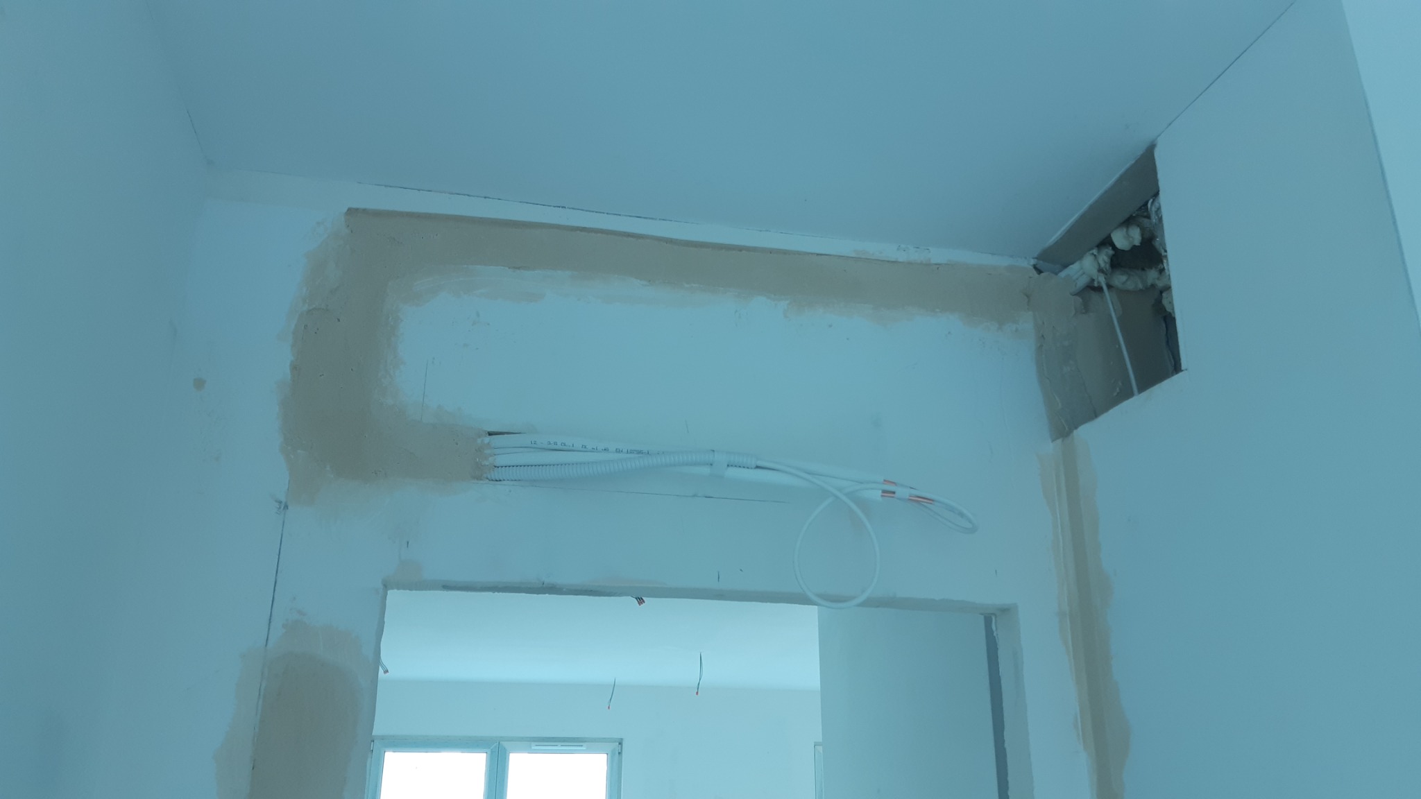 Białe kable elektryczne prowadzone w ścianie nad otworem drzwiowym, częściowo zakryte gipsem, z widoczną dziurą w rogu sufitu wypełnioną pianką montażową.