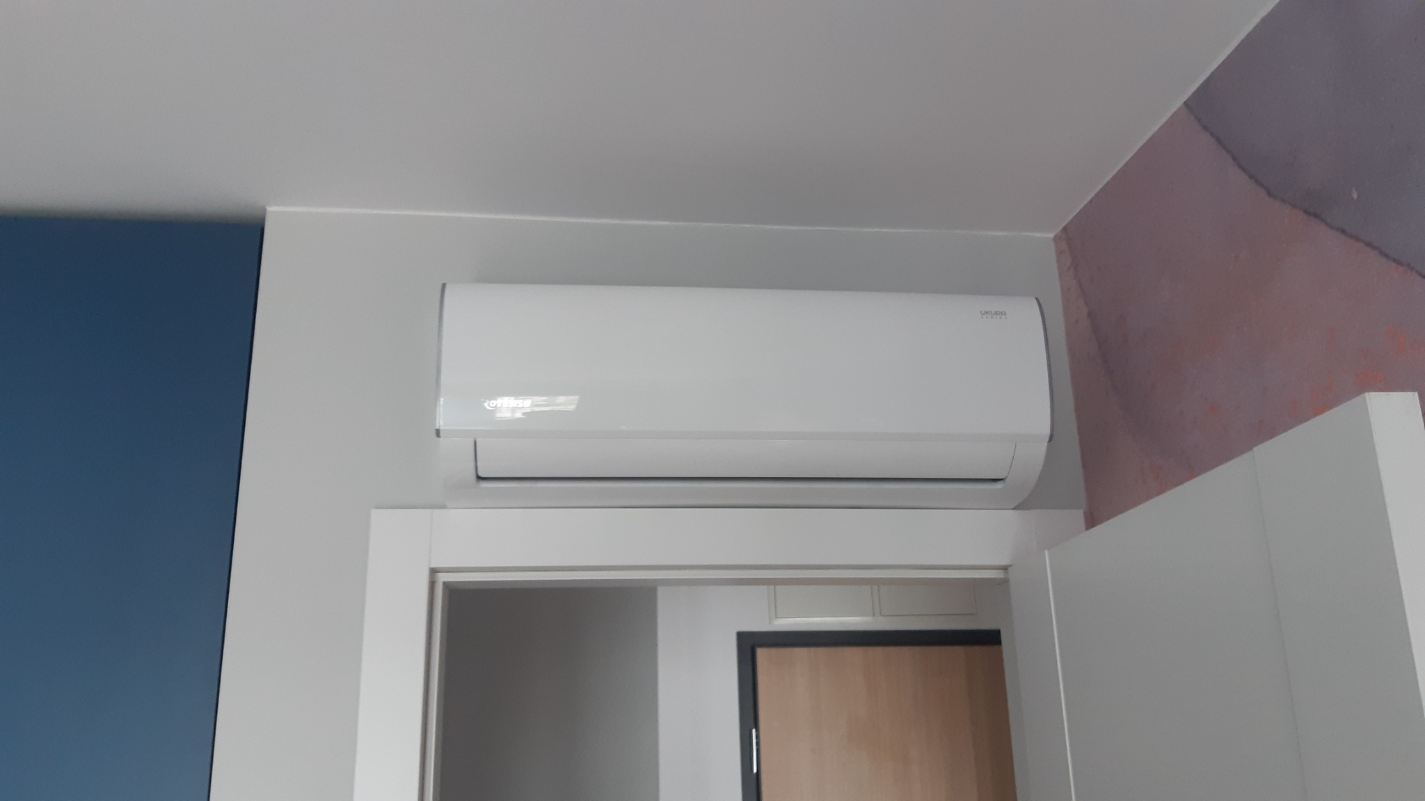 Biały klimatyzator marki Ukura zamontowany nad drzwiami w pomieszczeniu o niebieskiej i wzorzystej ścianie.