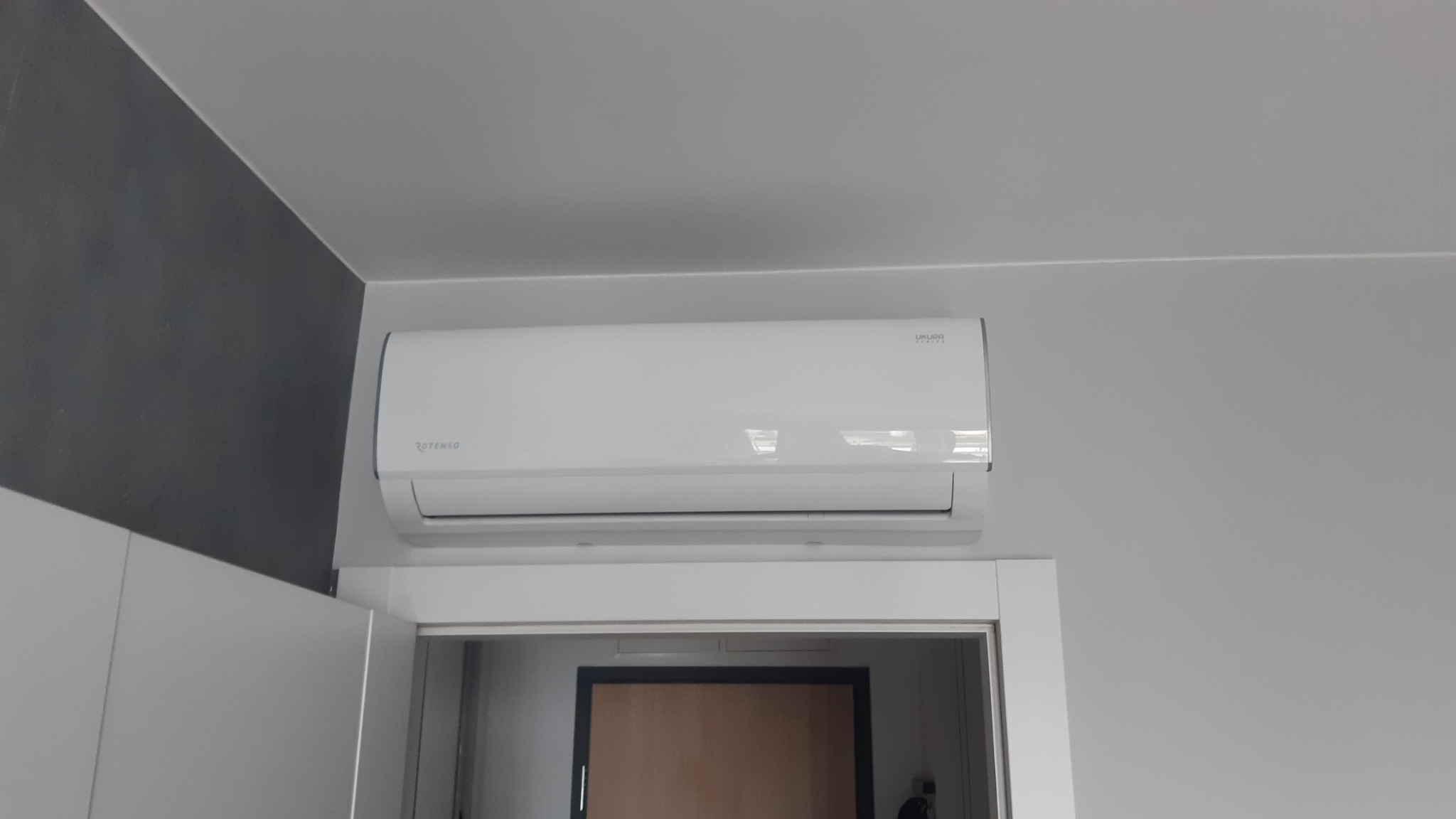 Biały klimatyzator marki Rotenso Ukura Series zamontowany nad futryną drzwi w jasnym pomieszczeniu z szarą ścianą.