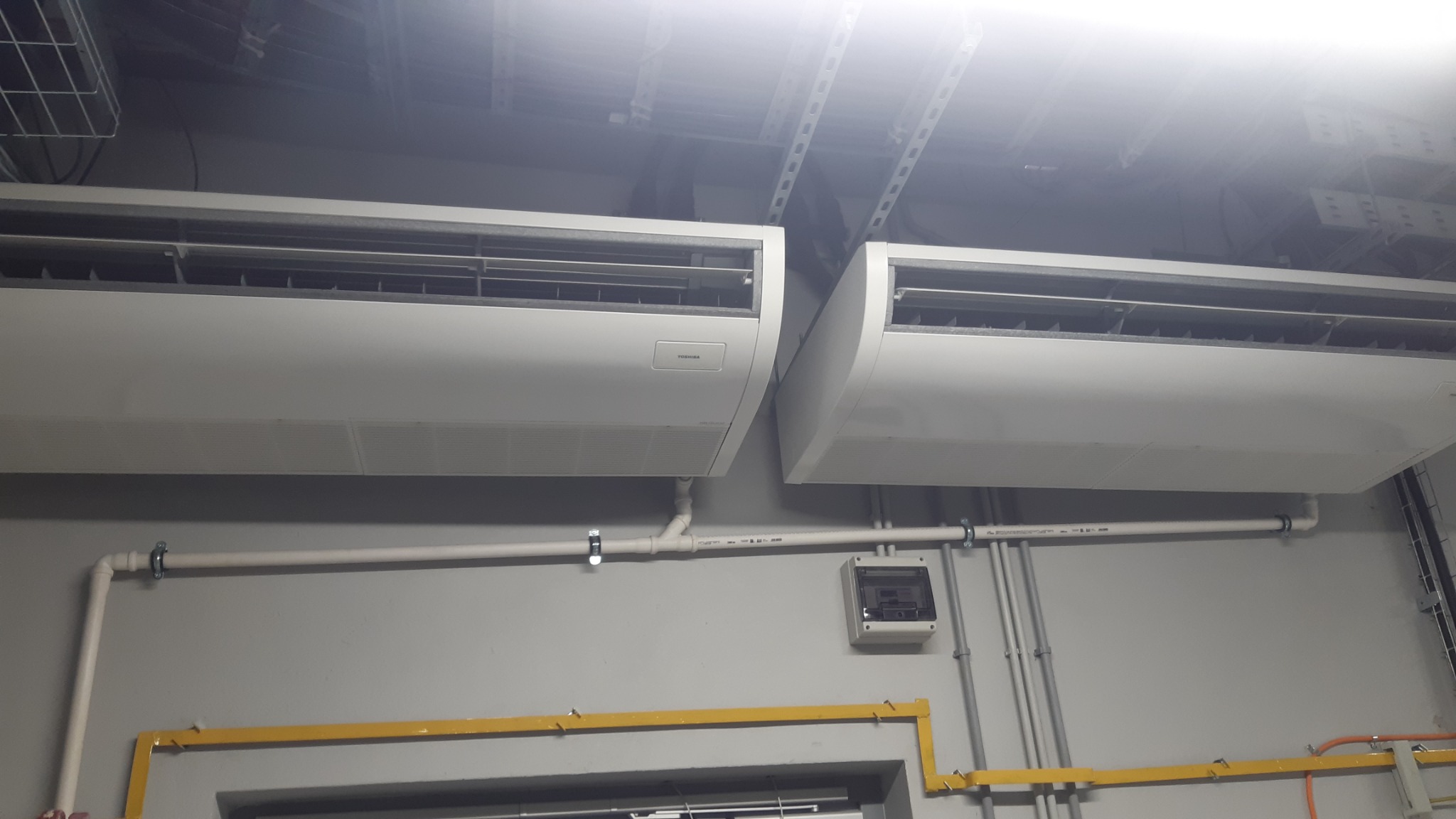 Dwa białe klimatyzatory Toshiba zamontowane pod sufitem z widocznymi rurami instalacyjnymi i skrzynką elektryczną na szarej ścianie.