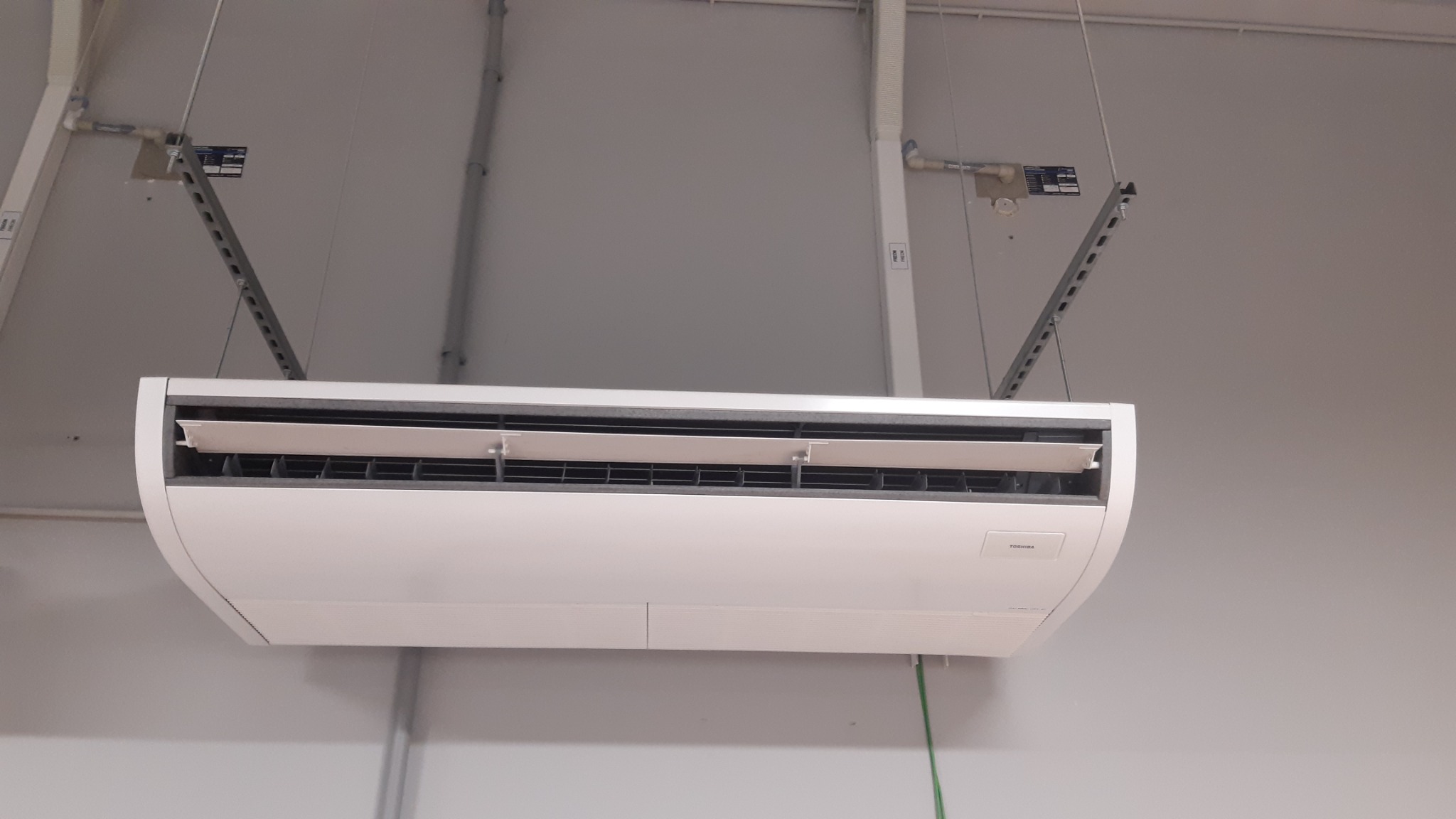 Biała jednostka klimatyzacyjna marki Toshiba zamontowana pod sufitem na metalowych wspornikach, widoczne przewody i instalacja sufitowa.