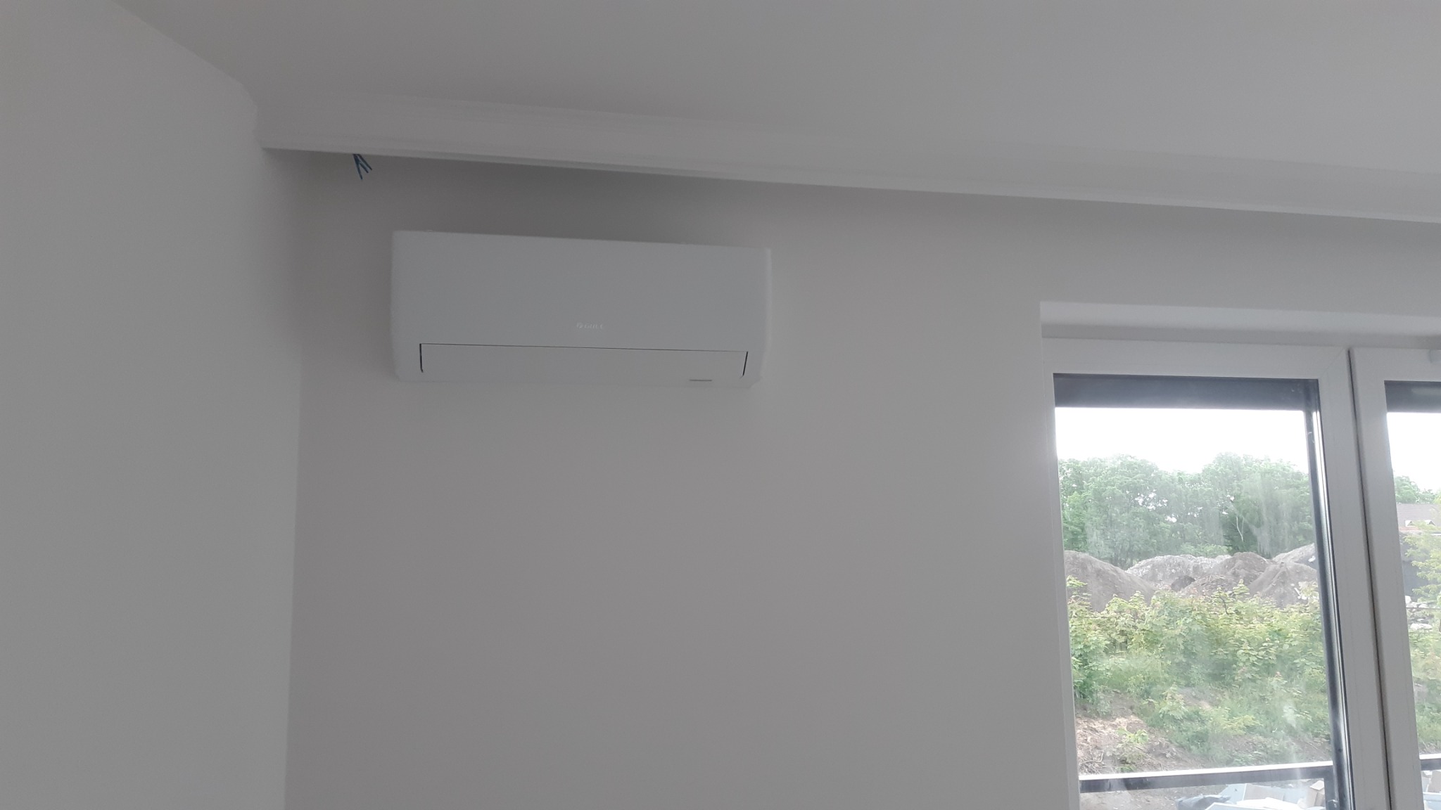 Biała jednostka wewnętrzna klimatyzacji zamontowana na ścianie w jasnym pomieszczeniu z widokiem na zewnątrz przez okno. Widoczne wystające kable spod listwy sufitowej.