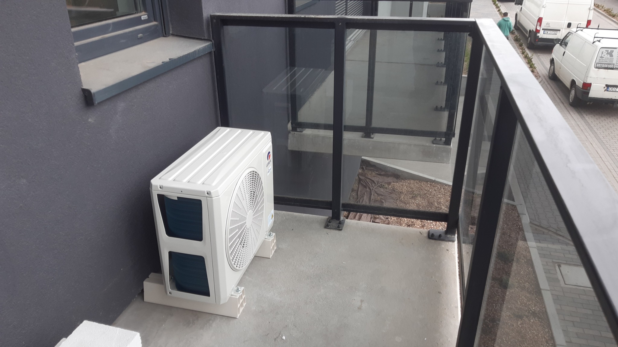 Biała jednostka zewnętrzna klimatyzacji marki Gree zamontowana na betonowym balkonie, na plastikowych podstawkach, z widoczną szarą elewacją budynku i szklaną balustradą z czarnymi elementami.