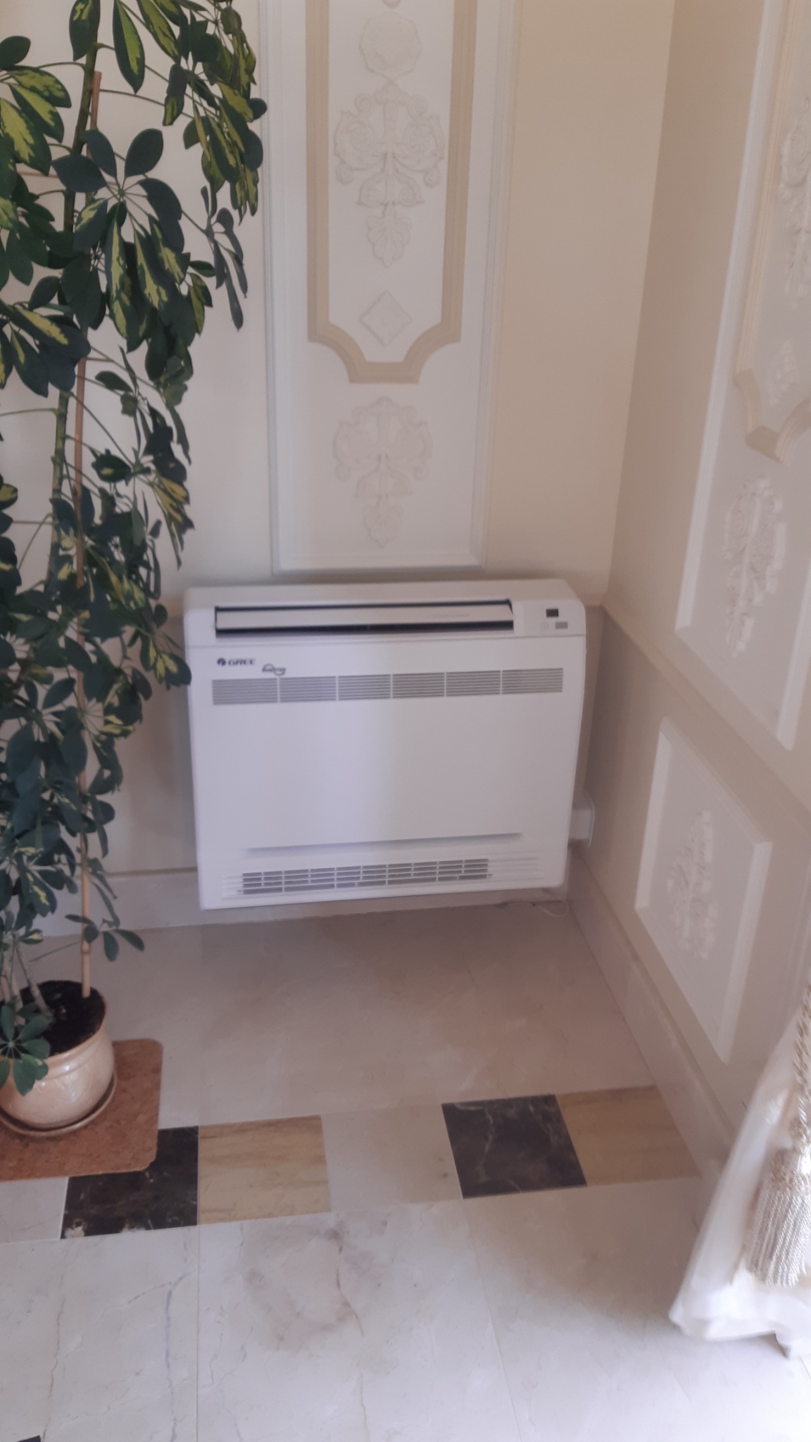 Biały klimatyzator podłogowy marki Gree w eleganckim wnętrzu z dekoracyjnymi panelami ściennymi i marmurową podłogą, obok doniczka z zieloną rośliną.