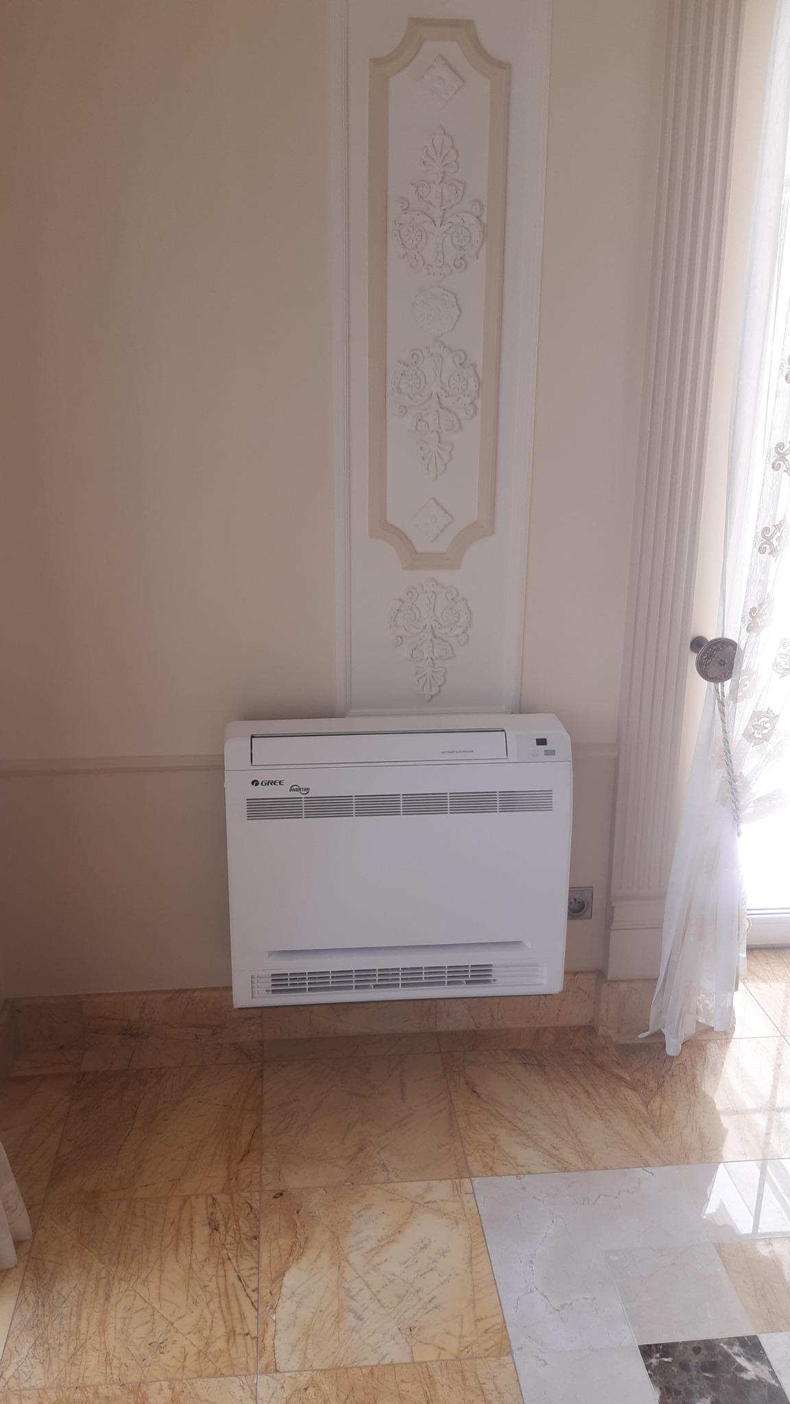 Biały klimatyzator podłogowy marki Gree zainstalowany w pomieszczeniu z dekoracyjną sztukaterią na ścianie i marmurową podłogą.