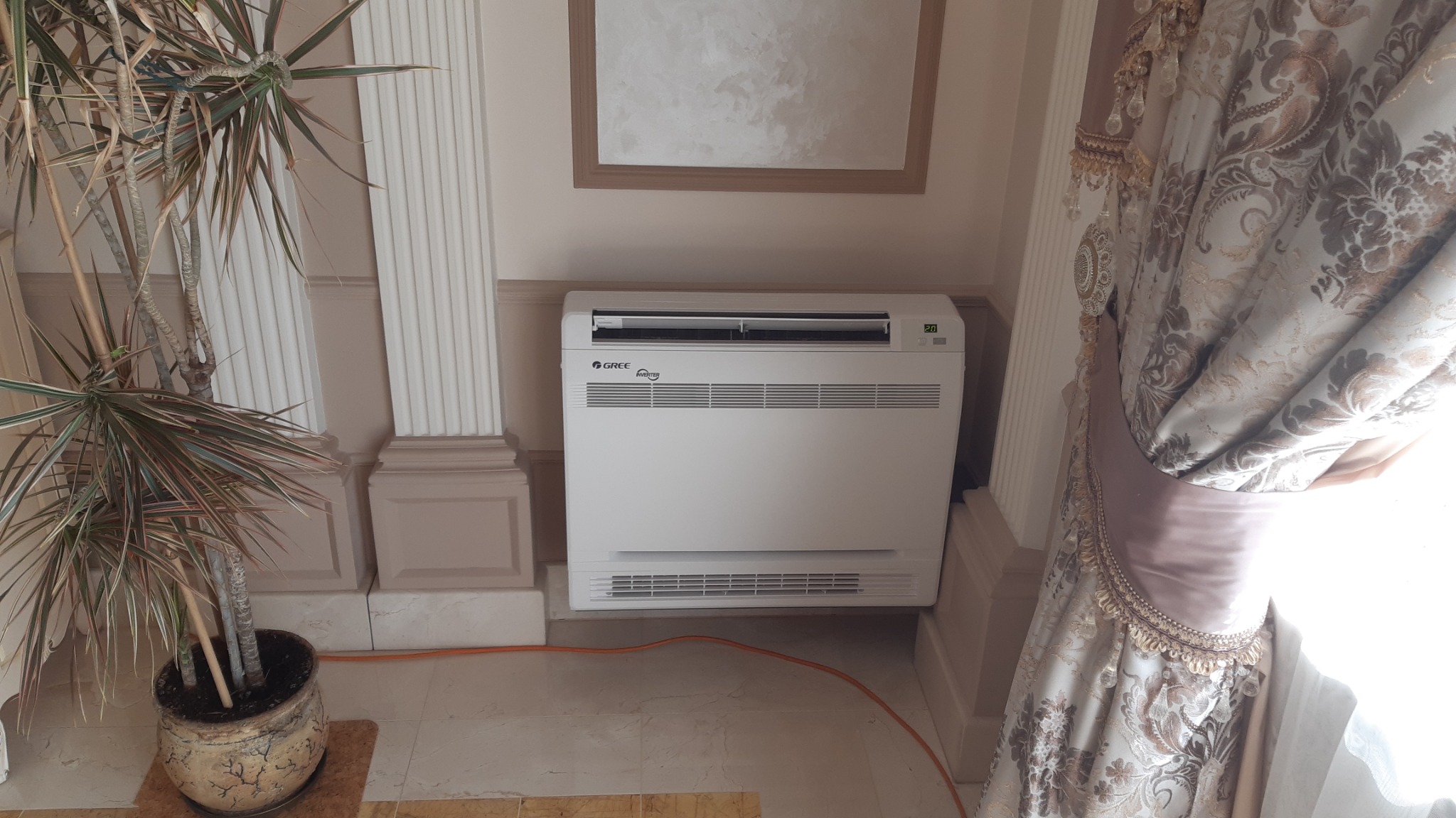 Biała jednostka wewnętrzna klimatyzacji pod oknem, widoczny fragment dekoracyjnej zasłony, donica z rośliną i dekoracyjne kolumny w tle.