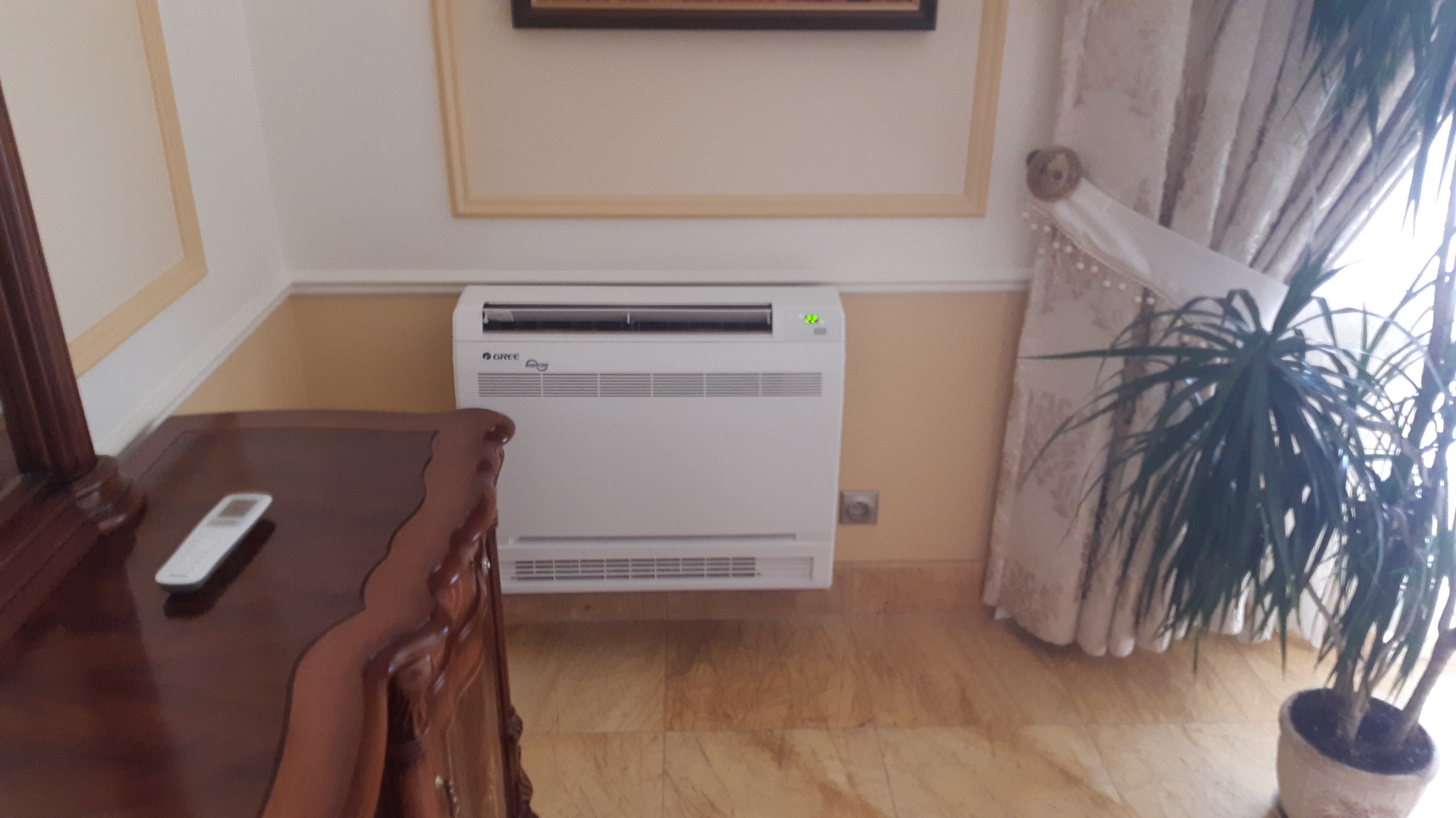 Biały klimatyzator podłogowy marki Gree w eleganckim salonie, z pilotem na drewnianej komodzie i rośliną doniczkową obok zasłony.