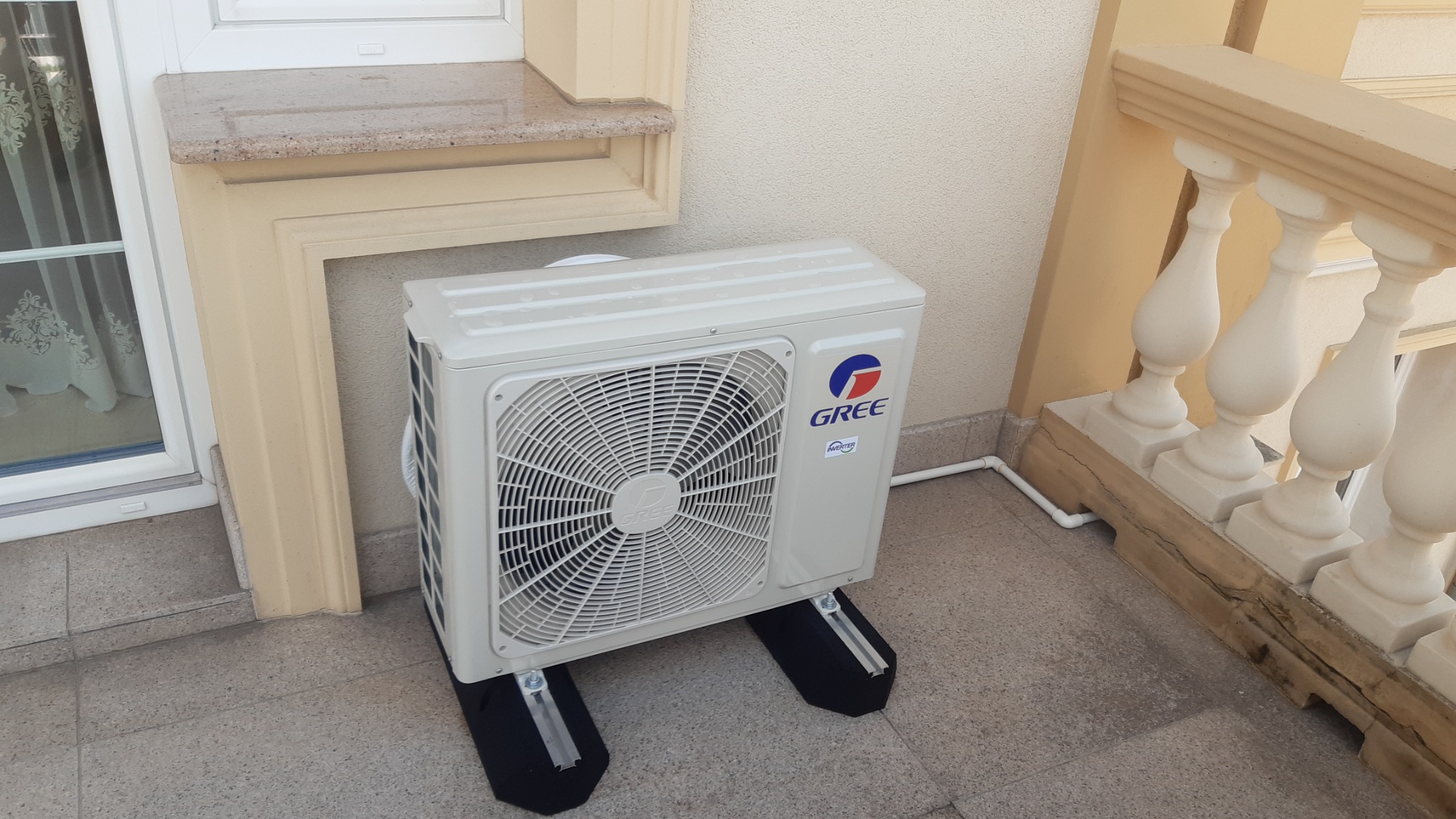 Biała jednostka zewnętrzna klimatyzatora marki Gree, umieszczona na czarnych podkładkach antywibracyjnych na balkonie z balustradą w stylu klasycznym.