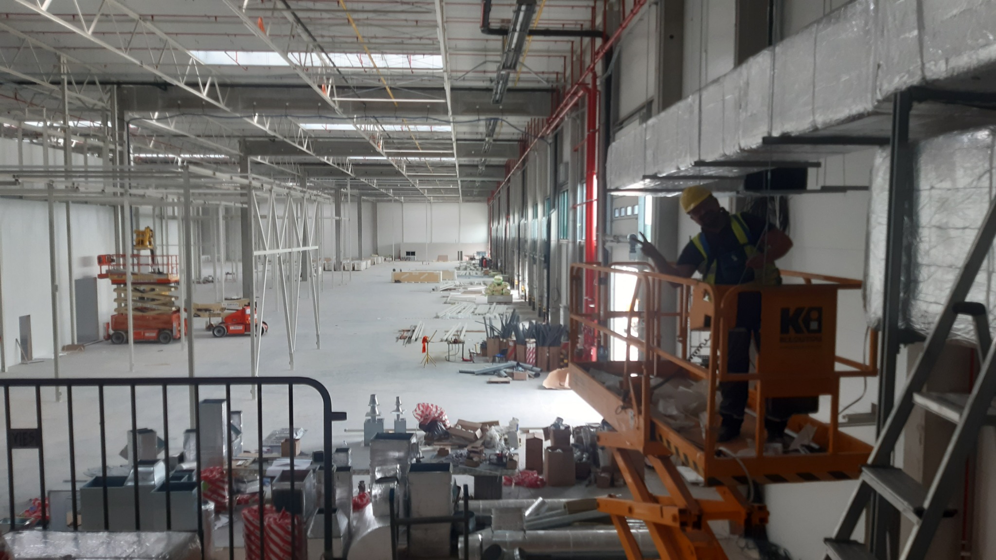 Pracownik w żółtym kasku na podnośniku koszowym montuje instalację wentylacyjną pod sufitem w hali przemysłowej z widocznymi elementami konstrukcyjnymi i przewodami wentylacyjnymi.