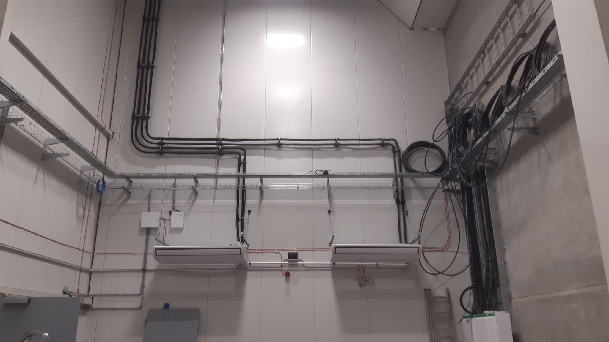 Dwa białe klimatyzatory zamontowane pod sufitem w pomieszczeniu przemysłowym, z widocznymi przewodami elektrycznymi i rurami chłodniczymi, prowadzącymi wzdłuż ściany i podwieszonymi na metalowych...