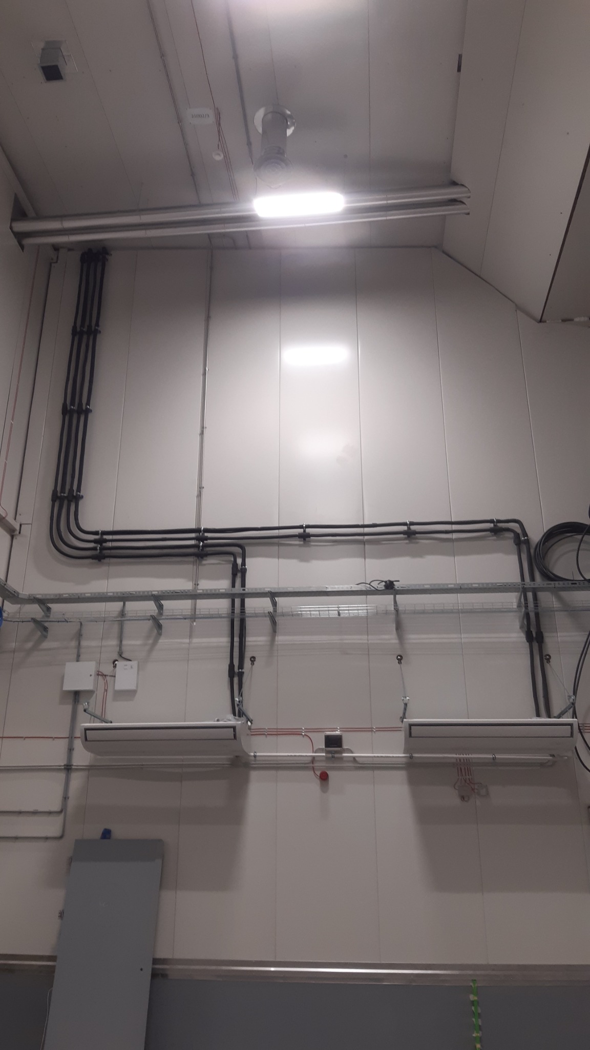 Wnętrze pomieszczenia przemysłowego z widocznymi instalacjami: rurami, przewodami elektrycznymi, korytami kablowymi oraz dwoma klimakonwektorami kasetonowymi przytwierdzonymi do ściany.