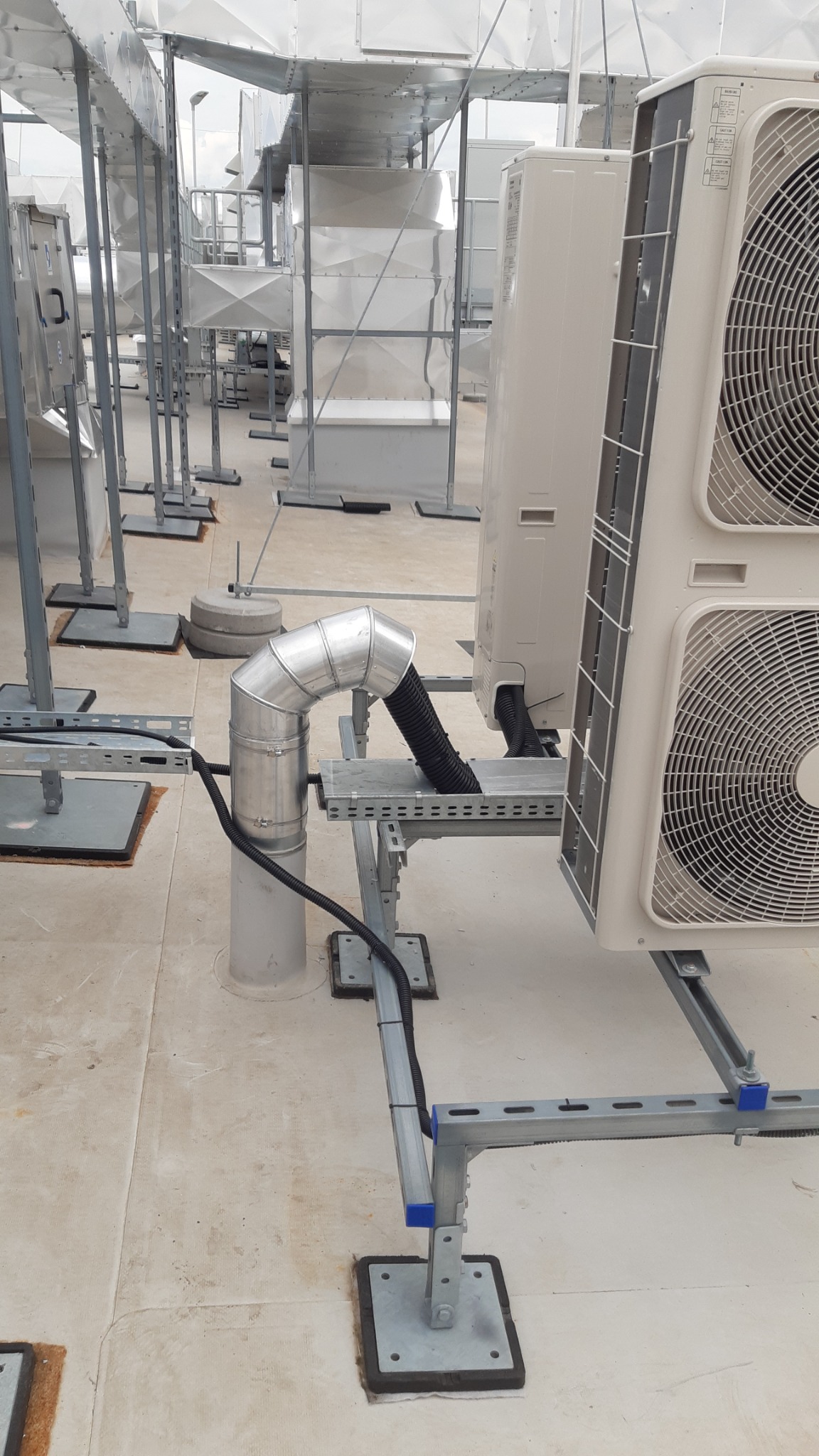 Zewnętrzne jednostki klimatyzacyjne zamontowane na dachu budynku komercyjnego, połączone metalowymi rurami i czarnym, karbowanym przewodem wentylacyjnym, wsparte na stalowej konstrukcji wsporczej.