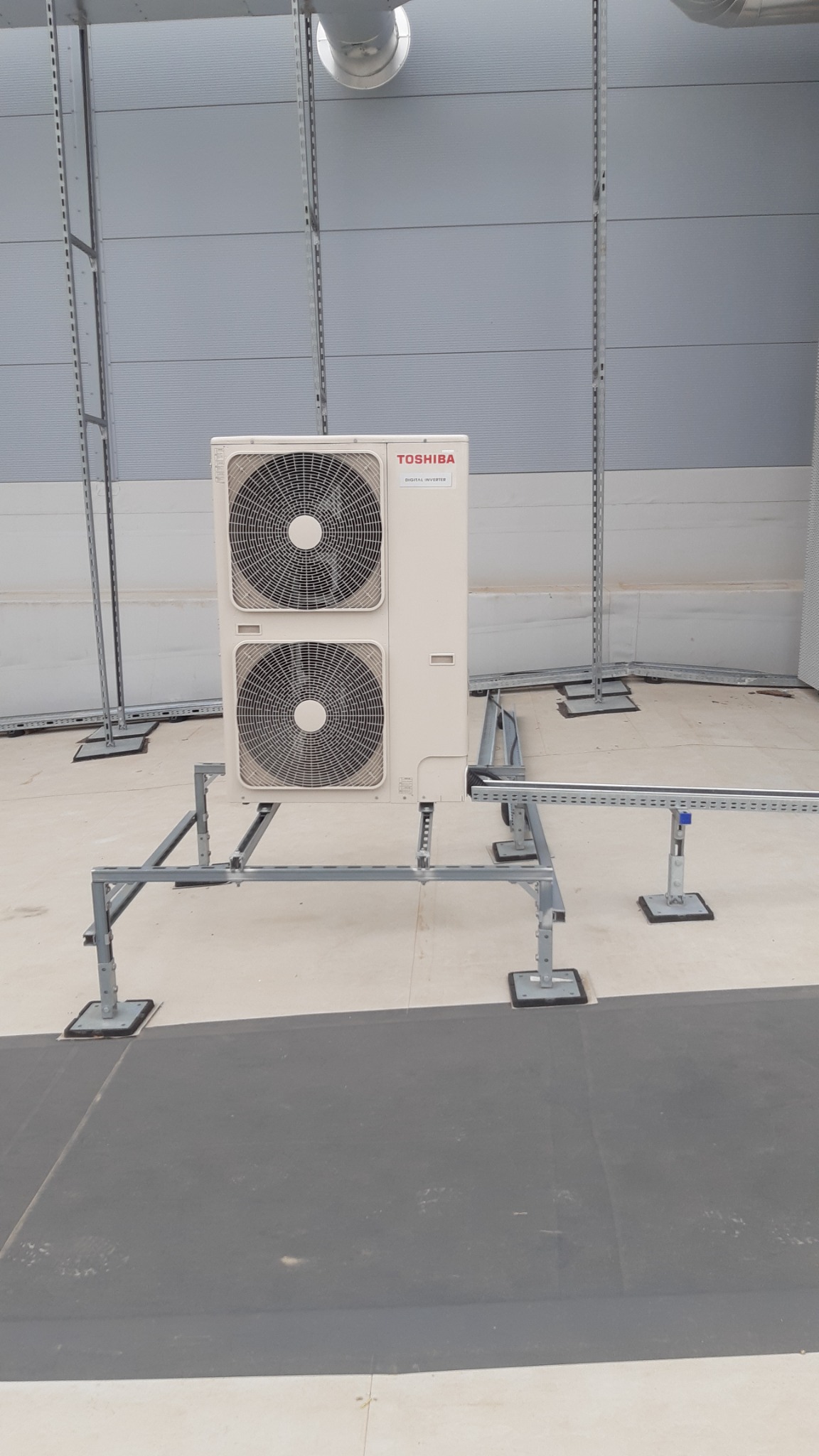 Zewnętrzna jednostka klimatyzacji Toshiba na metalowej konstrukcji wsporczej, zamontowana na dachu budynku przemysłowego, widoczne elementy wentylacji dachowej.