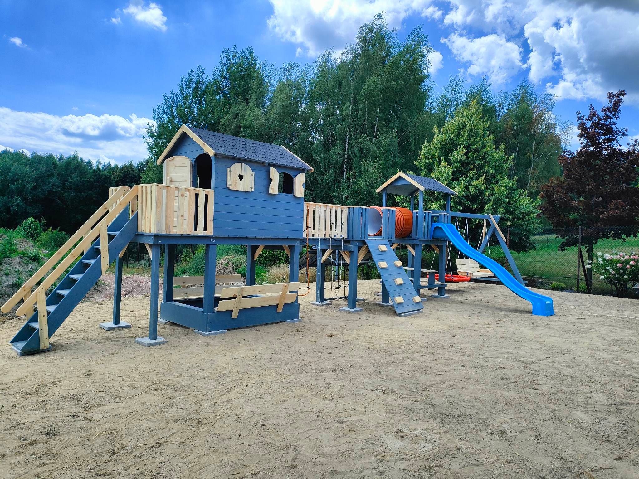 Kompleksowy plac zabaw w odcieniach szarości i naturalnego drewna, z domkiem na podeście, zjeżdżalnią i huśtawką, na piaszczystym podłożu, w tle drzewa i błękitne niebo z chmurami.