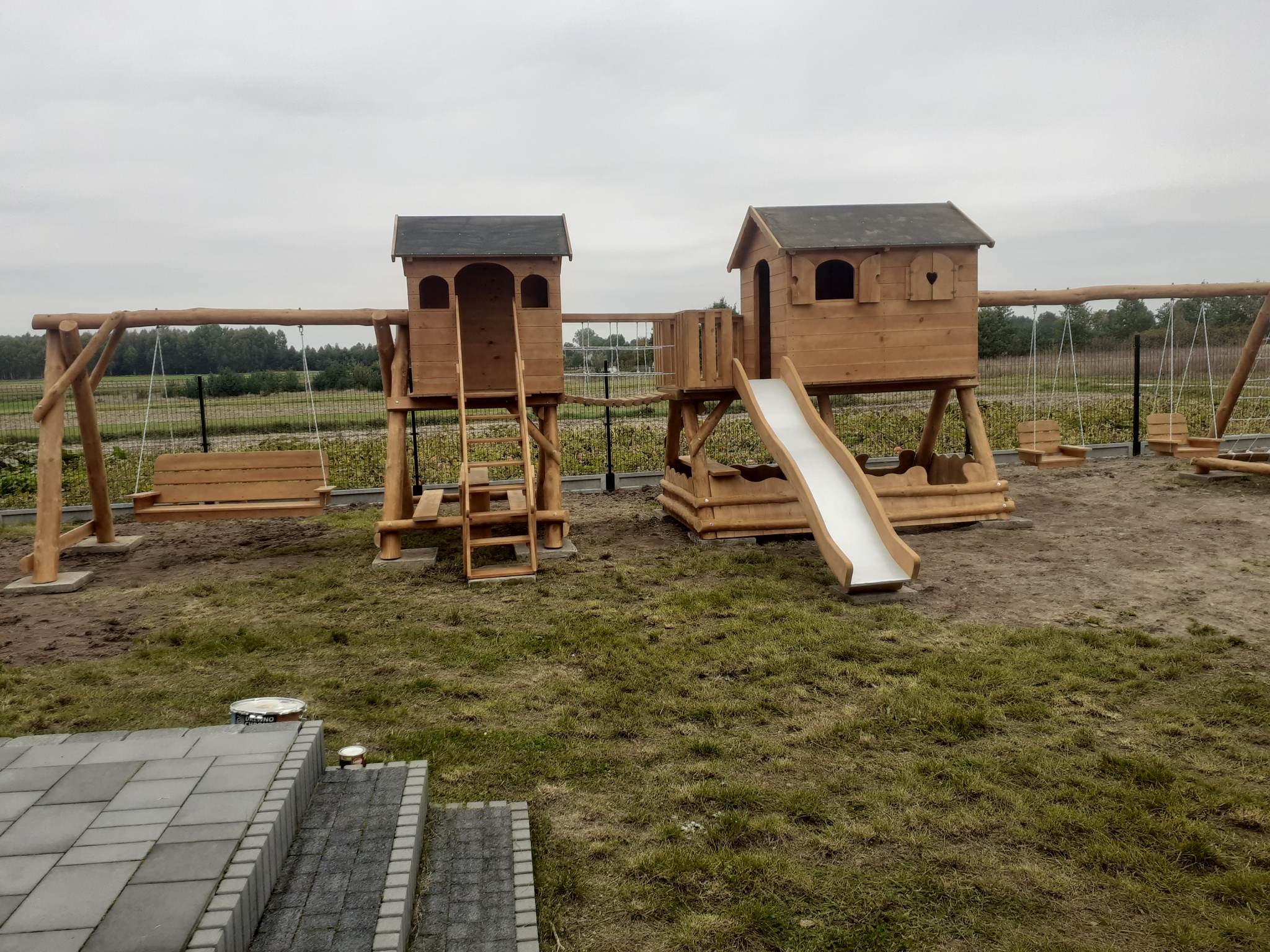 Drewniany plac zabaw z dwoma domkami na palach połączonymi mostkiem, huśtawkami i zjeżdżalnią, widok z poziomu ziemi, puszki z impregnatem na pierwszym planie.