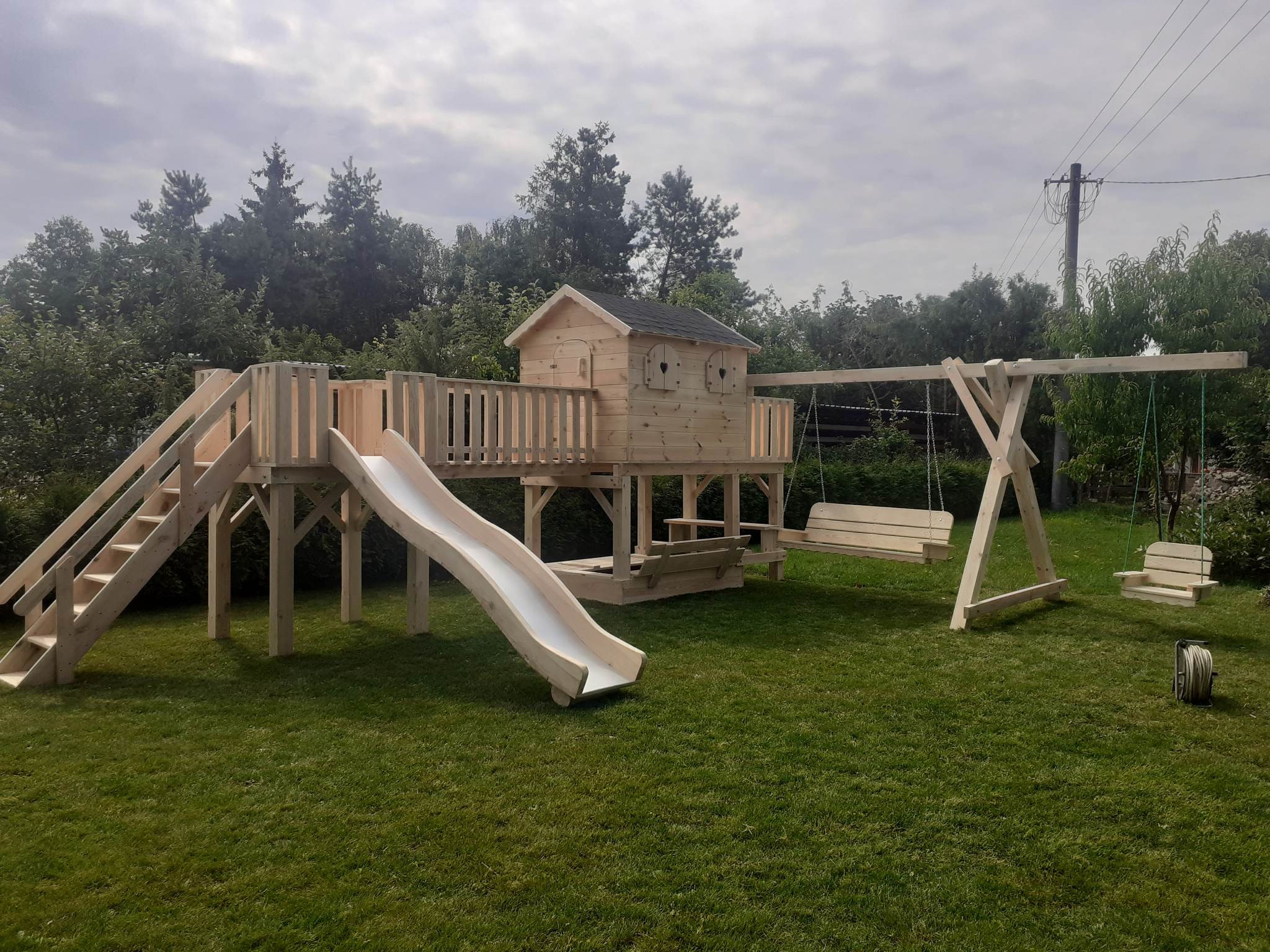 Drewniany plac zabaw z domkiem na podeście, zjeżdżalnią i huśtawkami na tle zielonego trawnika i drzew