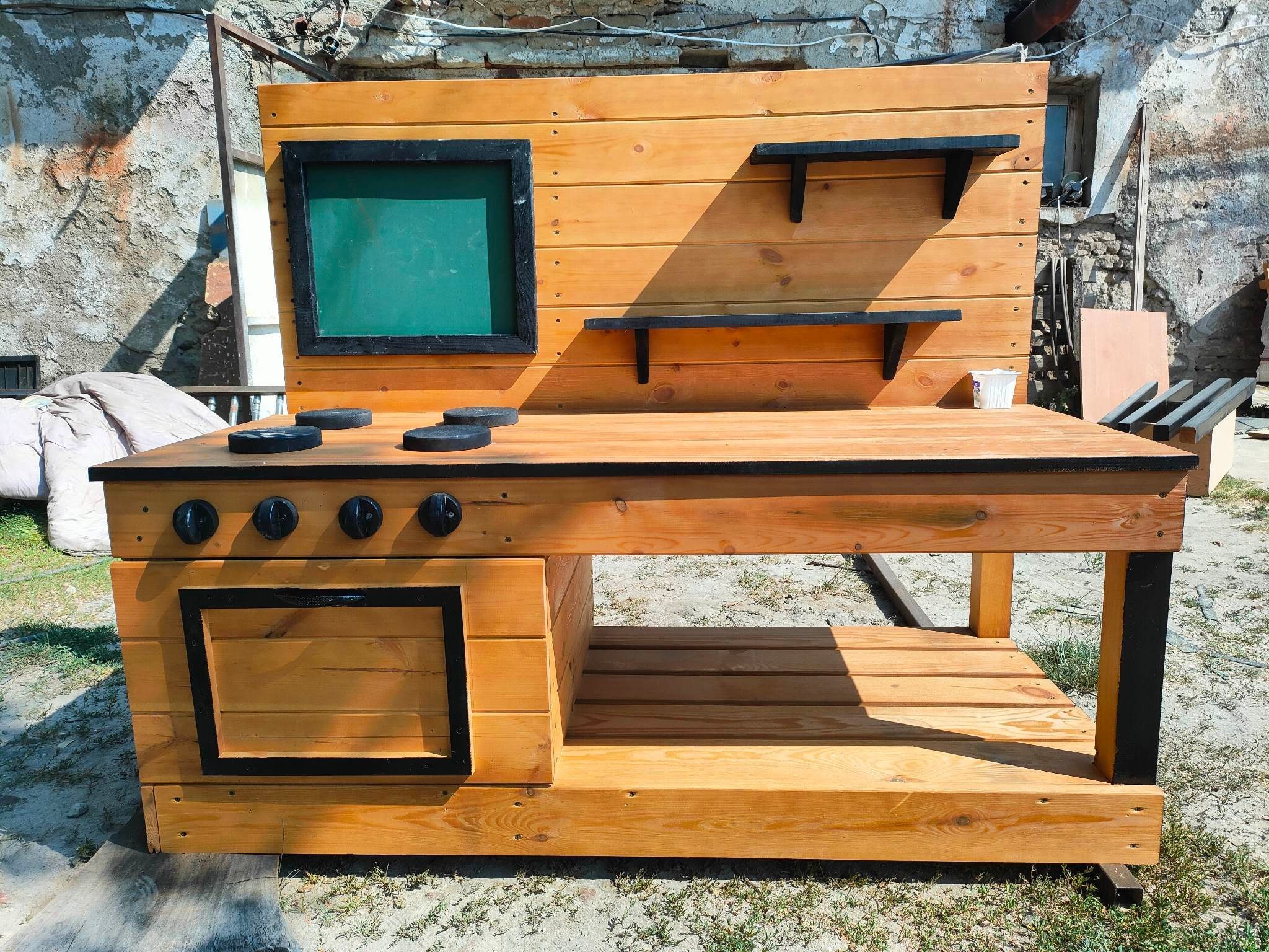 Drewniana kuchnia do zabawy na zewnątrz z blatem imitującym kuchenkę, piekarnikiem i tablicą kredową, ustawiona na piaszczystym podłożu z widocznym murem w tle.
