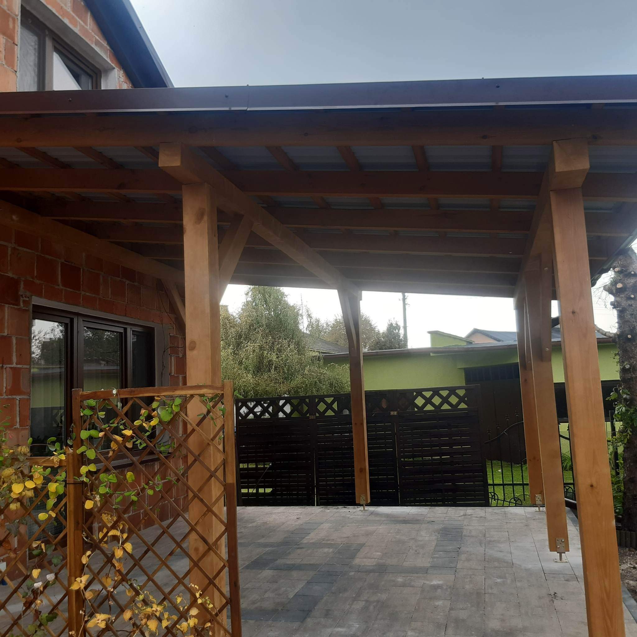Drewniana wiata z metalowym dachem, widoczna z zewnątrz, z ażurową ścianką z pnączami, na podłożu z kostki brukowej.