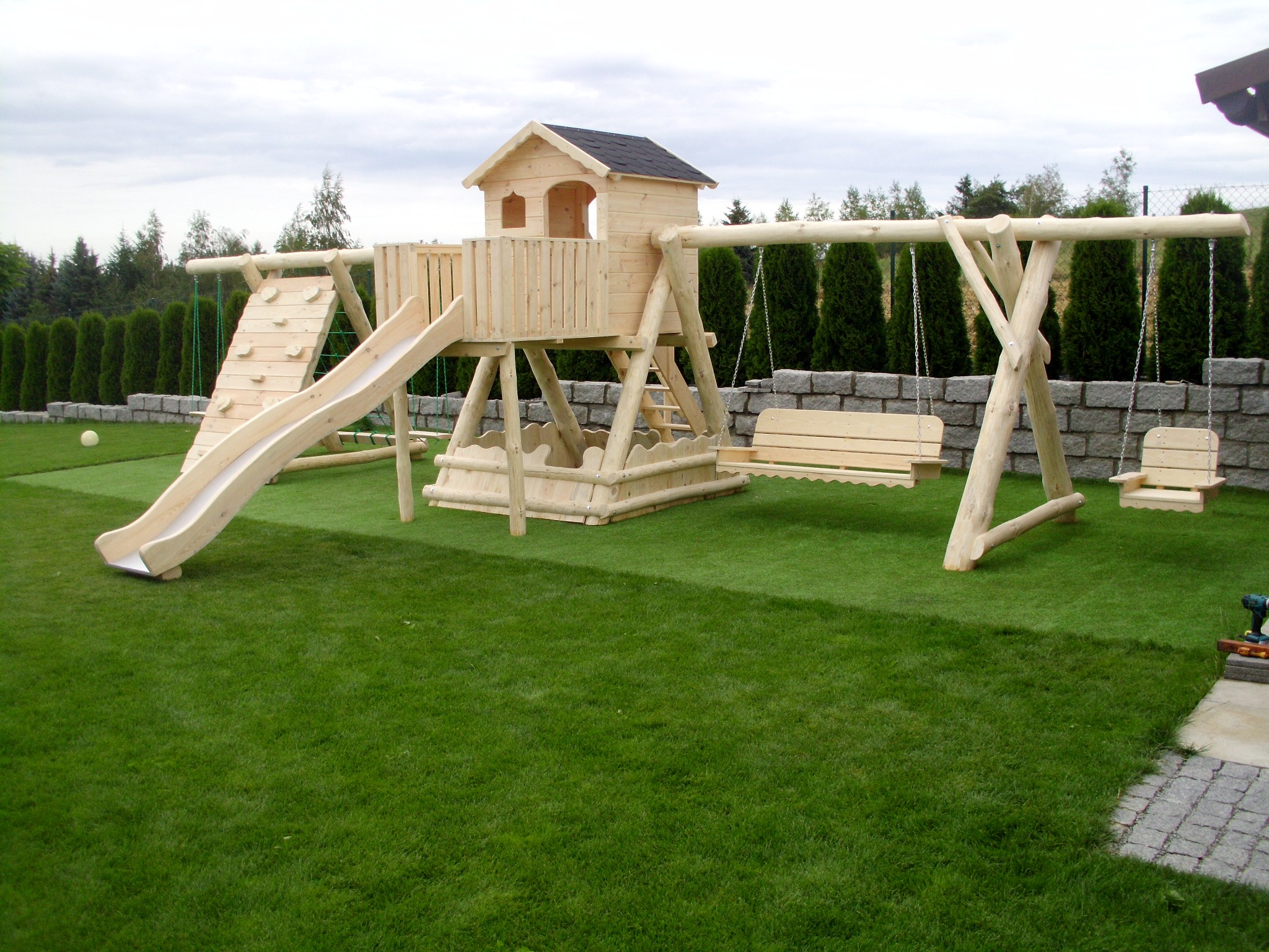 Drewniany plac zabaw z domkiem, zjeżdżalnią i huśtawkami na tle zielonego trawnika i żywopłotu; w prawym dolnym rogu widać wkrętarkę.