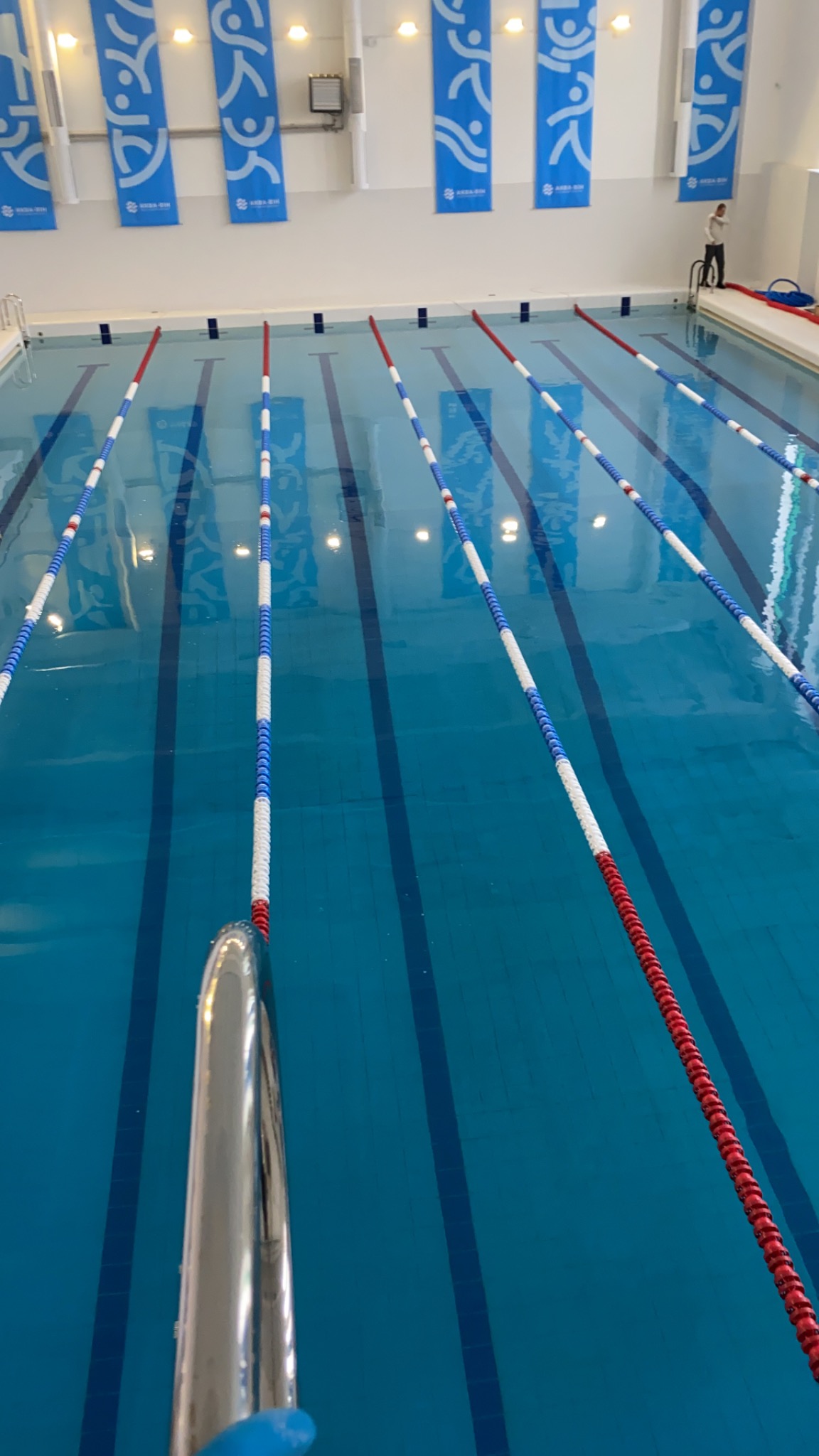 Kryty basen sportowy z torami oddzielonymi linami w kolorach czerwonym, białym i niebieskim, widok z perspektywy skraju basenu.