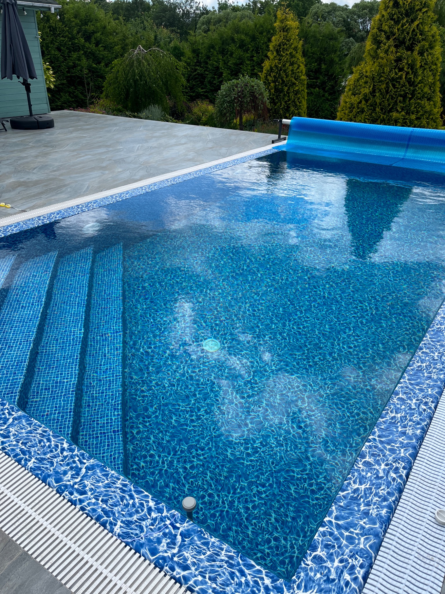 Błękitny basen z mozaikową wykończeniem, widoczne schody wejściowe i częściowo zwinięta pokrywa, otoczony zielenią.