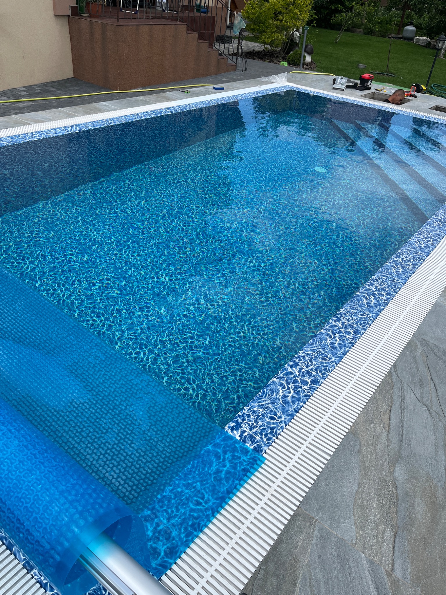 Błękitny basen z mozaiką wykończony szarym kamieniem, częściowo zakryty niebieską folią ochronną.