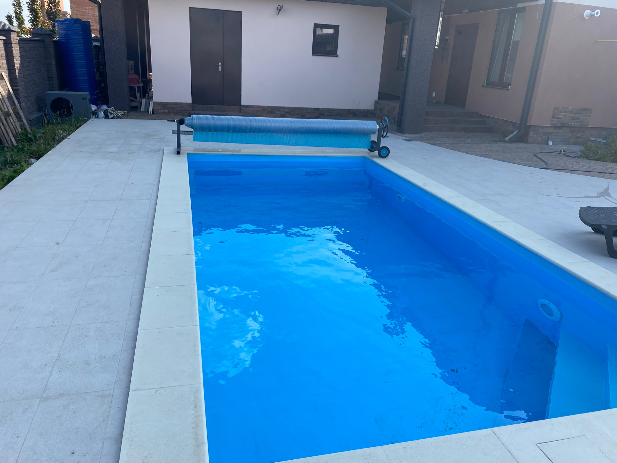 Błękitny basen ogrodowy z automatyczną roletą zabezpieczającą, otoczony jasnymi płytami chodnikowymi, na tle budynku z ciemnymi drzwiami i oknami.