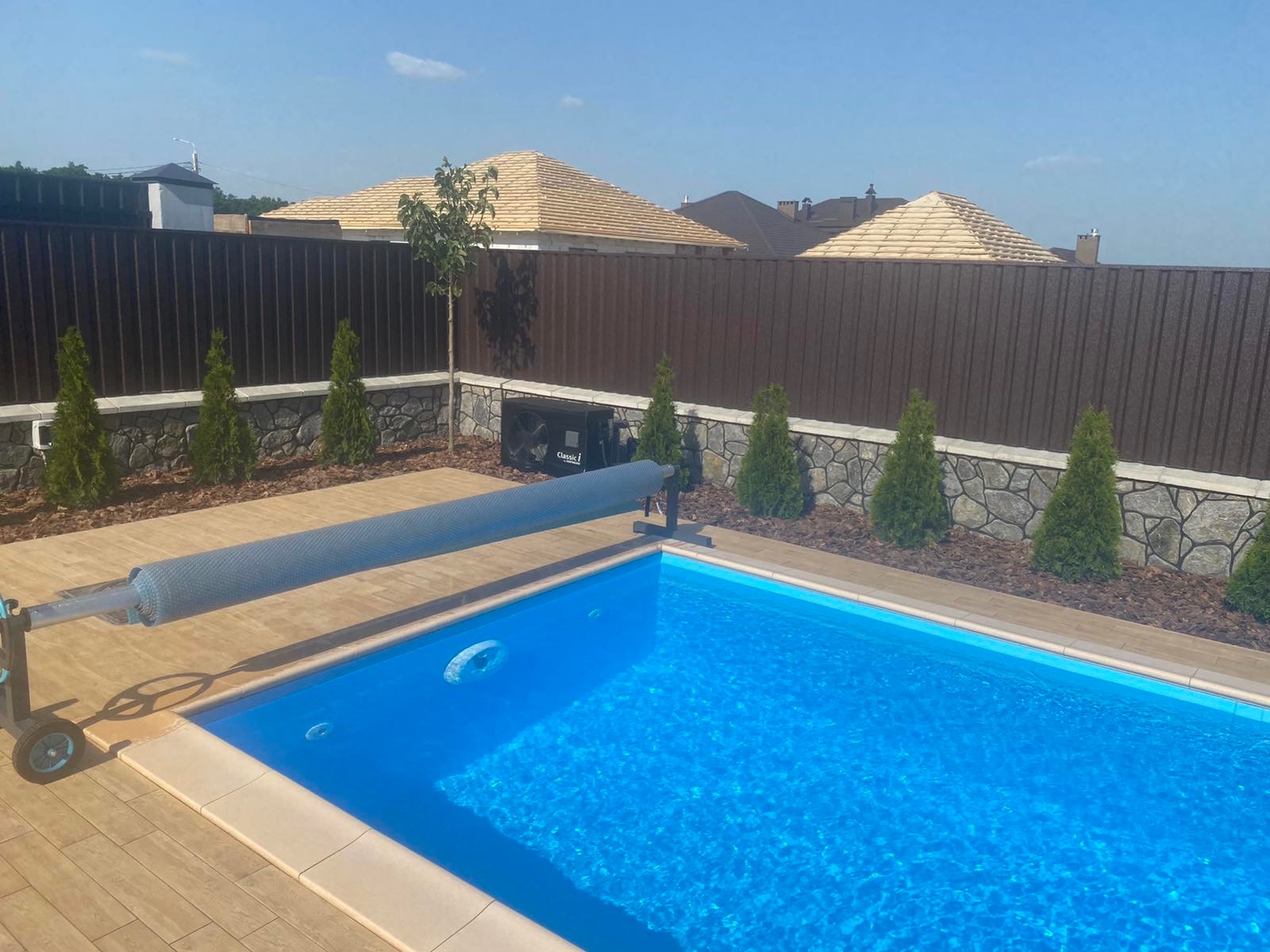 Prostokątny basen z błękitną wodą, częściowo zakryty szarą roletą na rolce, otoczony drewnianym tarasem, z widocznym systemem ogrzewania wody, tujami i brązowym ogrodzeniem.