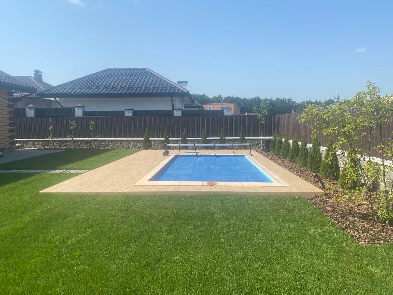 Zewnętrzny basen z niebieskim przykryciem, otoczony drewnianym tarasem i zadbanym trawnikiem, w tle ogrodzenie i budynek mieszkalny.