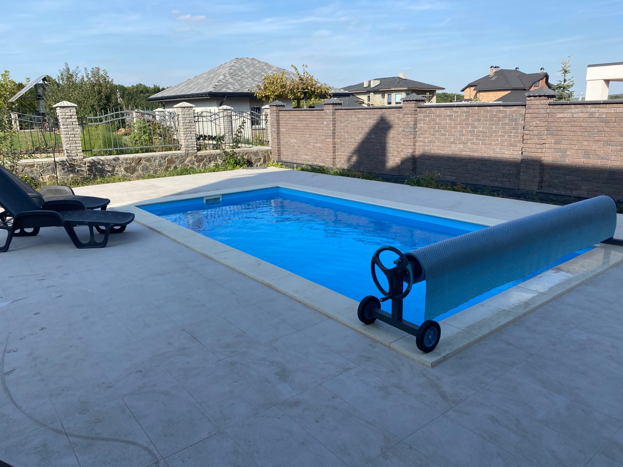 Prostokątny basen z błękitną wodą i zwiniętą szarą pokrywą na rolce, otoczony jasnymi płytkami, w tle domy z dachami i ceglany mur.