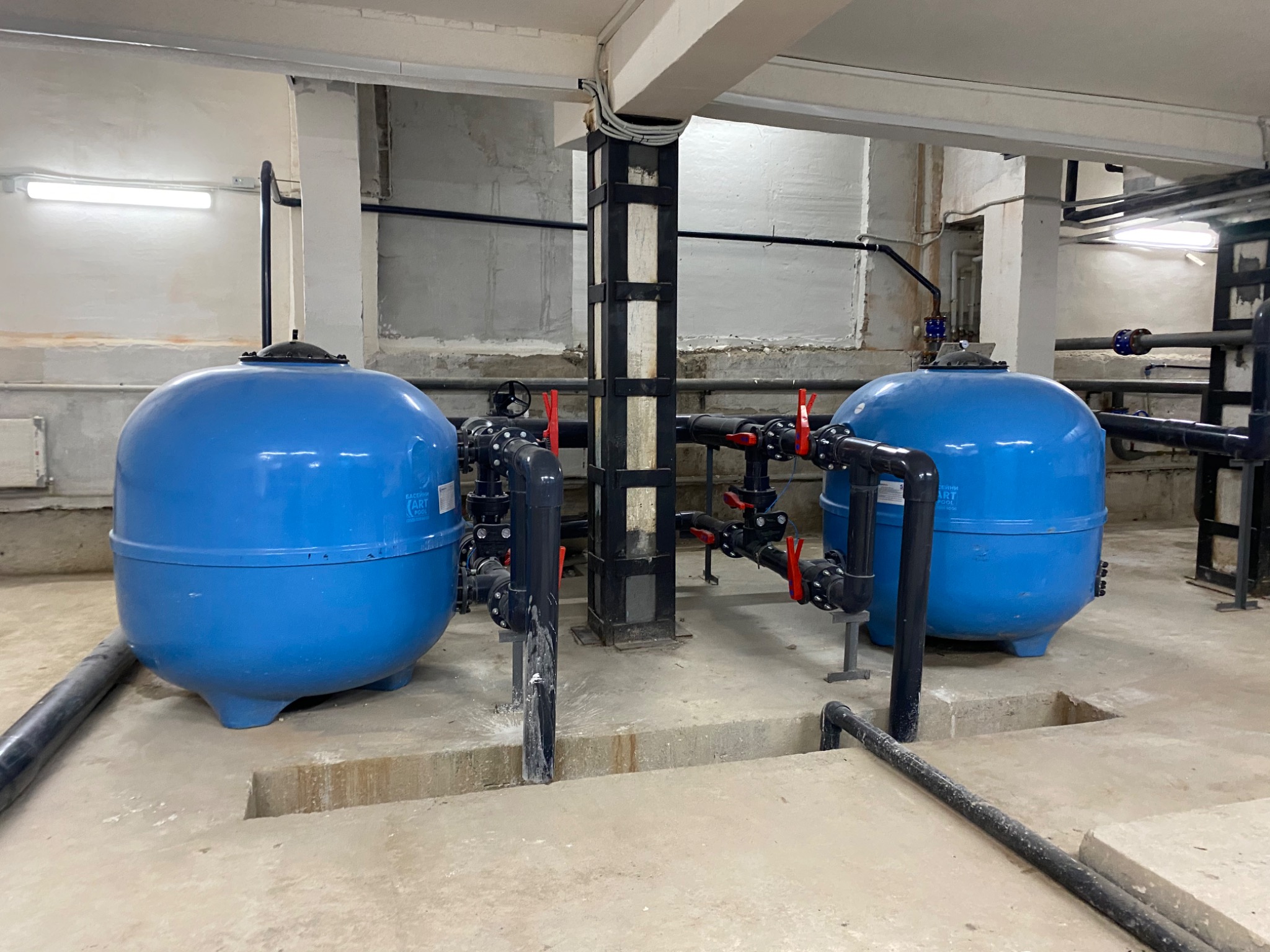 Dwa niebieskie filtry basenowe w pomieszczeniu technicznym z instalacją rur i zaworów, widoczne betonowe wnęki w podłodze.