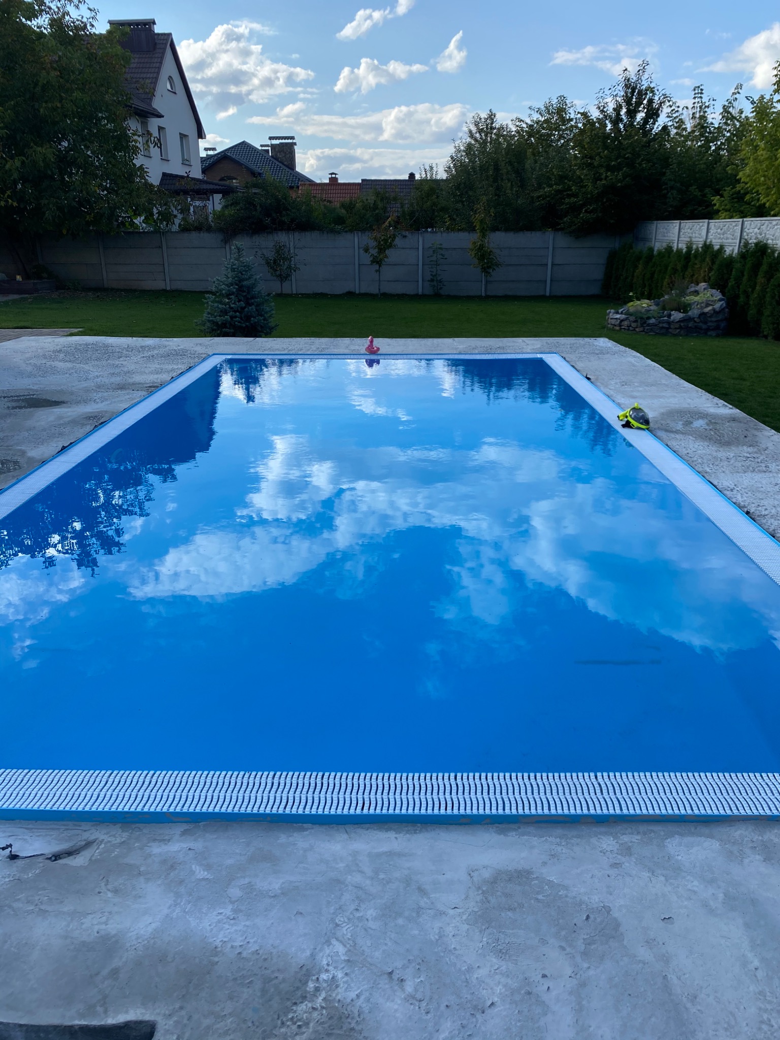 Napełniony basen ogrodowy z błękitną wodą odbijającą chmury, na brzegu leży maska do nurkowania, w tle dom i zielony trawnik.