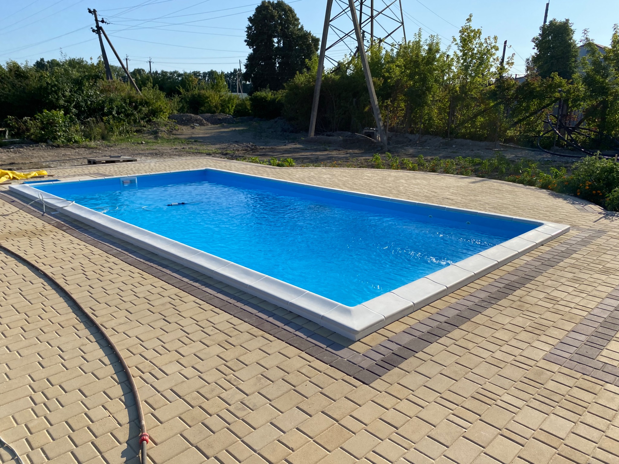 Prostokątny basen z błękitną wodą, otoczony jasną kostką brukową z ciemniejszym pasem obramowania, widok z góry, w tle słupy wysokiego napięcia i zieleń