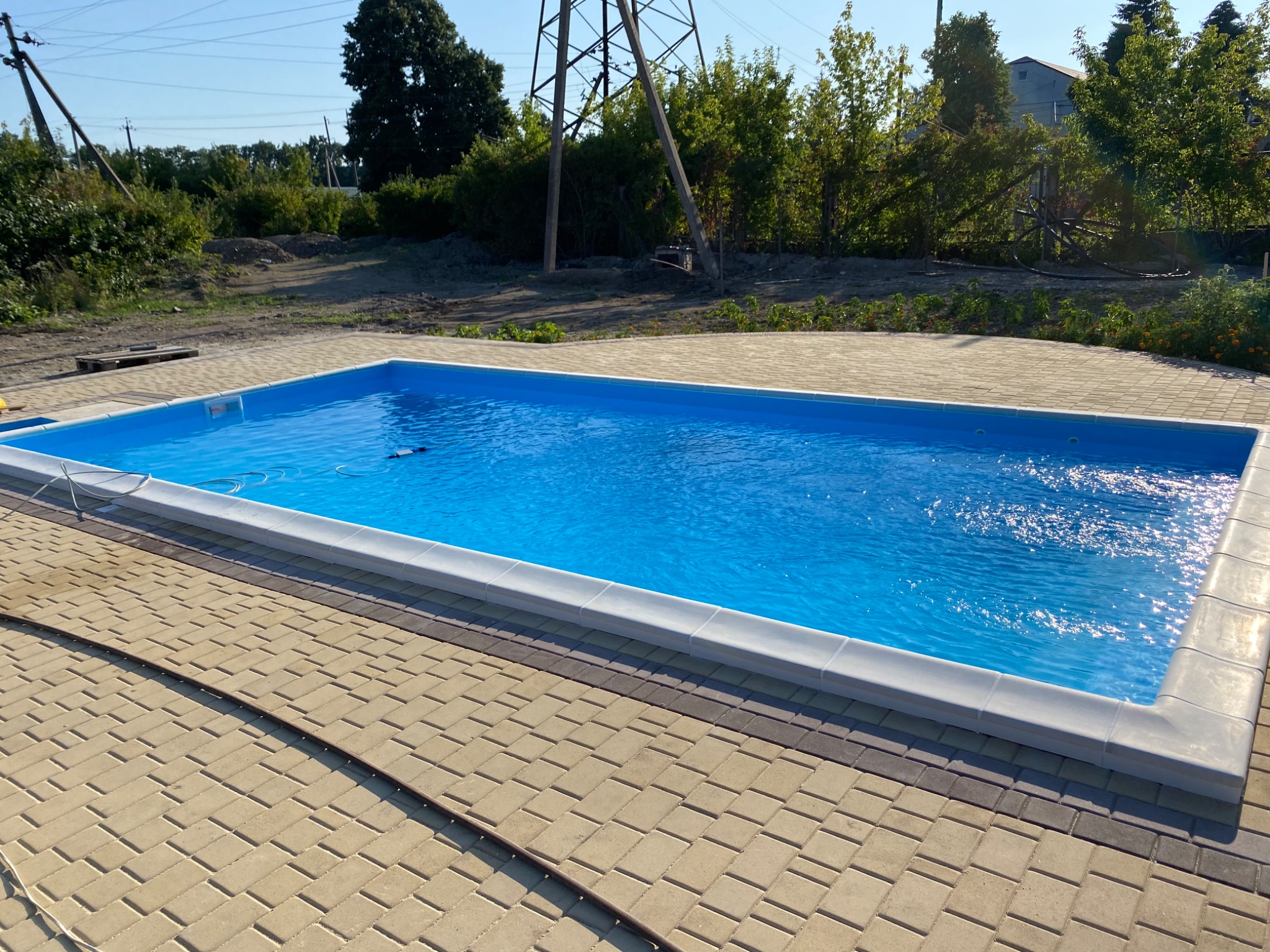 Prostokątny basen z błękitną wodą, otoczony jasną kostką brukową, na tle zieleni i słupów energetycznych.