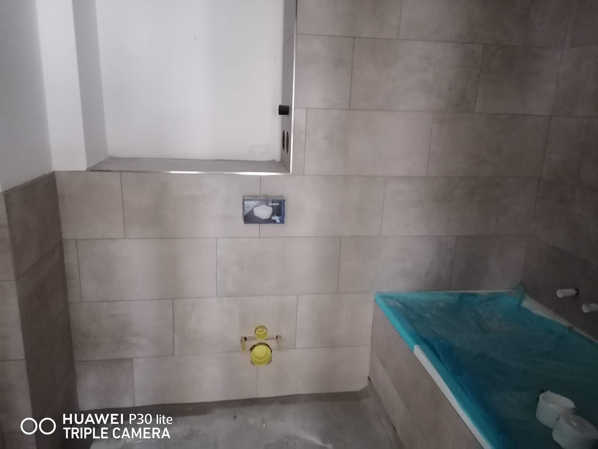 Montaż płytek ściennych w łazience z widoczną instalacją podtynkową do WC i osłoniętą folią wanną.