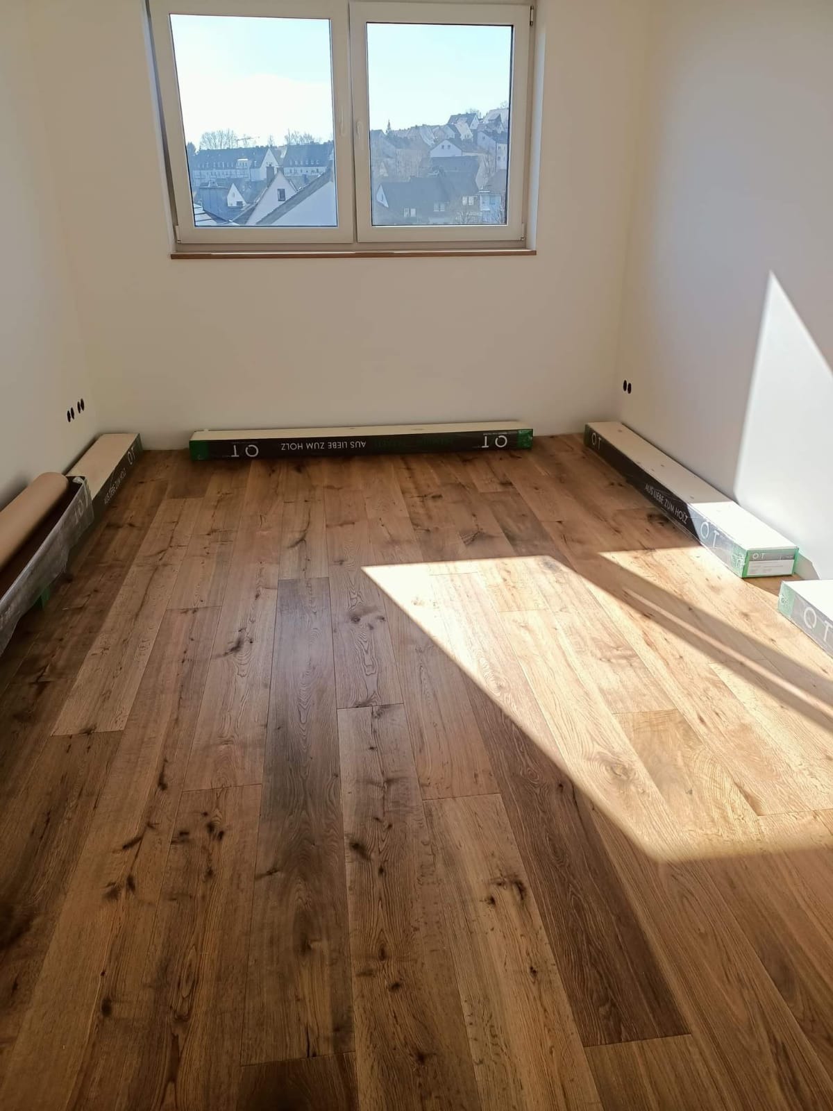 Nowo położona, jasna podłoga drewniana w pustym pokoju z oknem i listwami przypodłogowymi, widoczne opakowania paneli podłogowych przy ścianach.