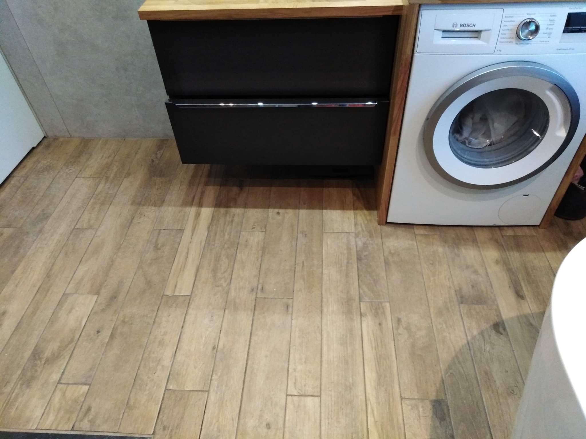 Nowoczesna łazienka z drewnopodobną podłogą, czarną szafką pod umywalkę z drewnianym blatem i pralką marki Bosch.