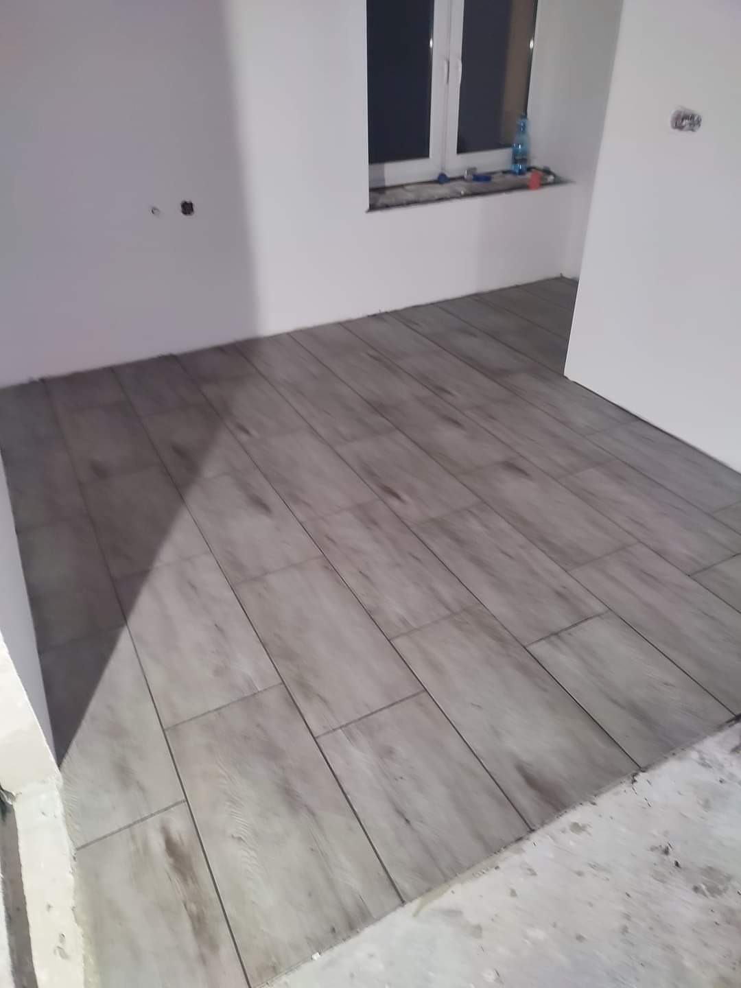 Ułożona podłoga z płytek imitujących drewno w jasnym odcieniu szarości, widoczne ściany w trakcie prac wykończeniowych, okno z butelką wody na parapecie.