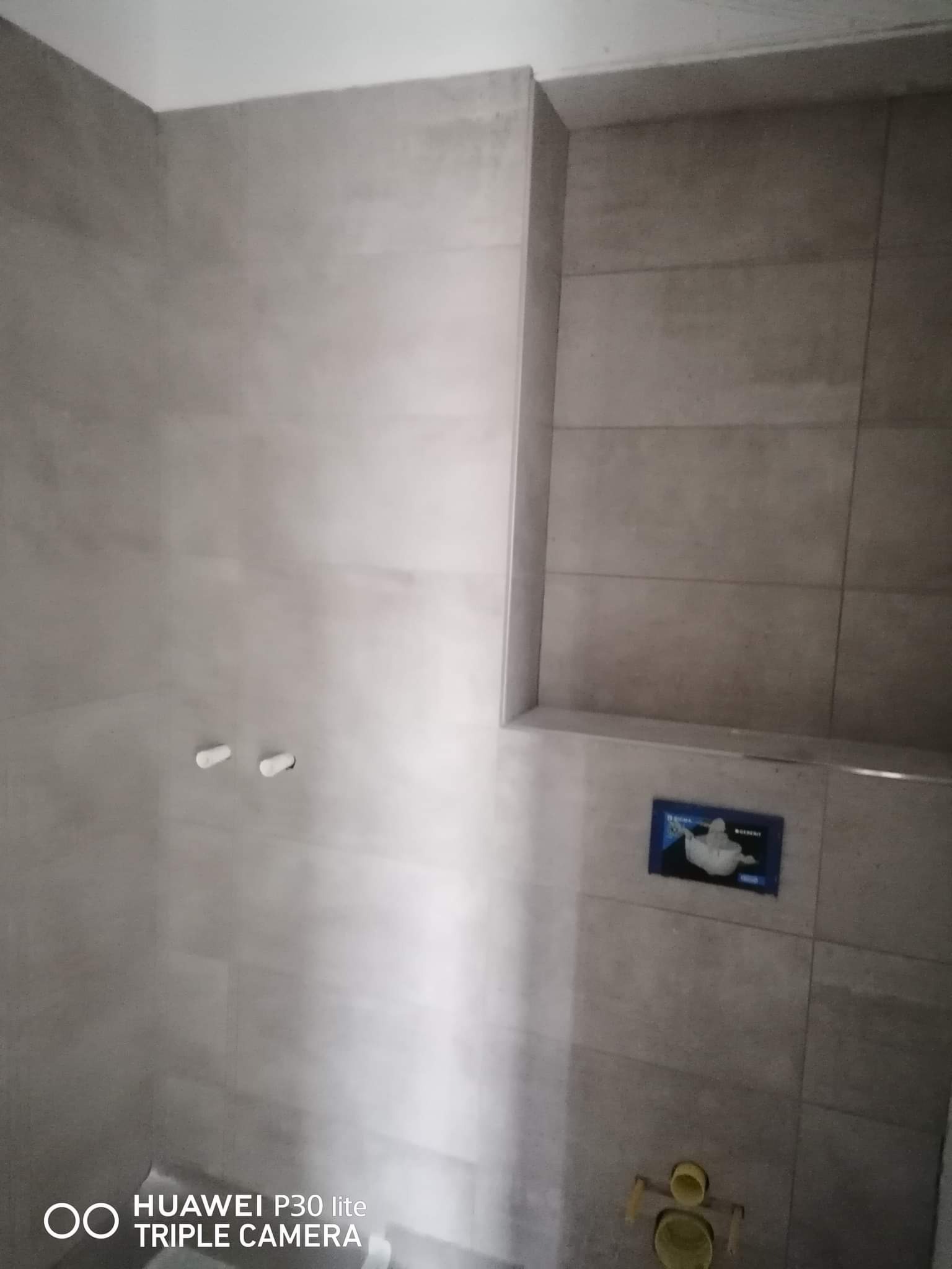 Szare płytki ceramiczne ułożone na ścianach łazienki, widoczne przyłącza do armatury i zabudowany stelaż podtynkowy WC z niebieską instrukcją montażu.